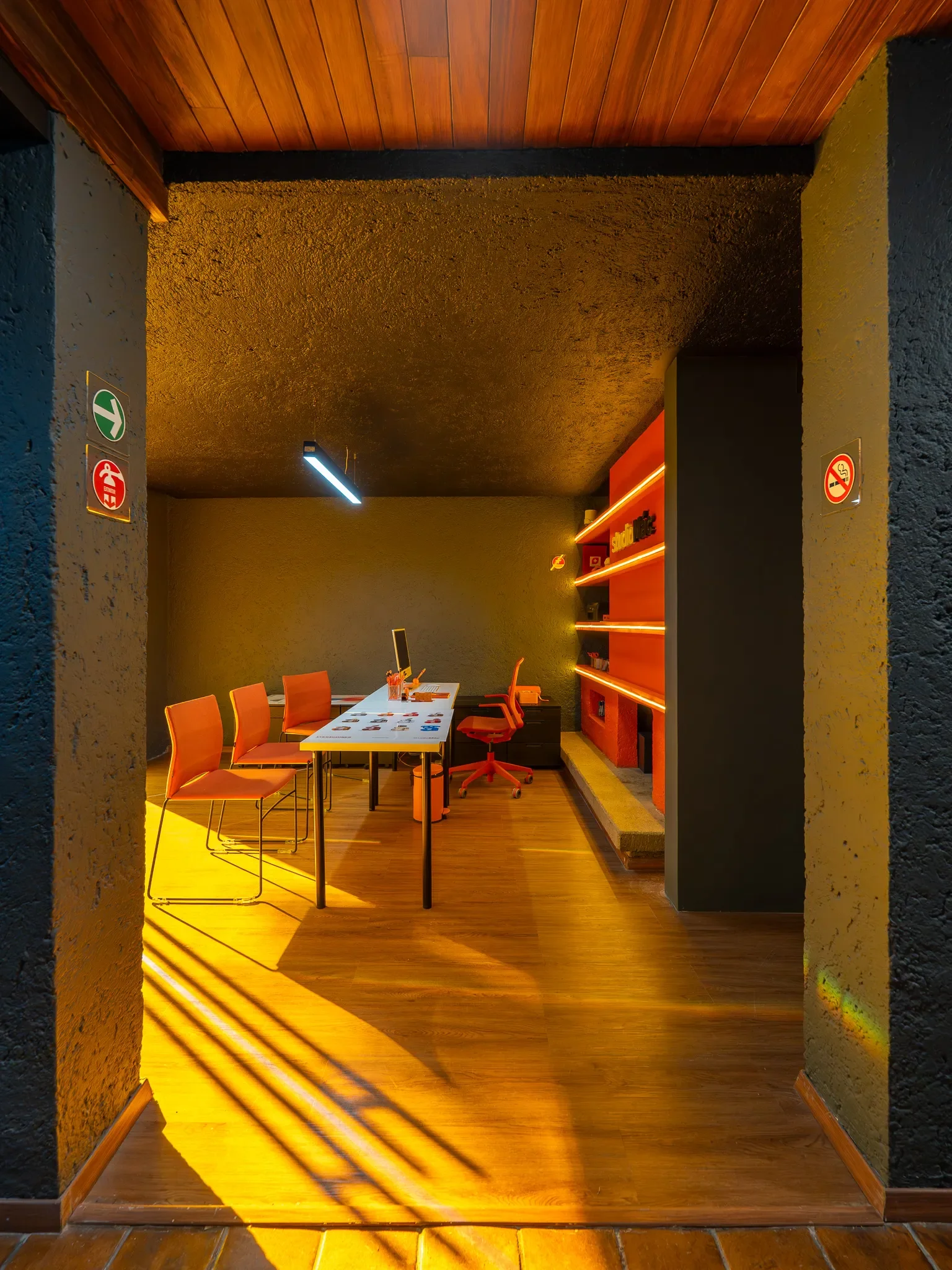 Sala de reuniones moderna con mesa blanca y sillas naranjas, estantería roja con decoraciones y señalamiento de no fumar.