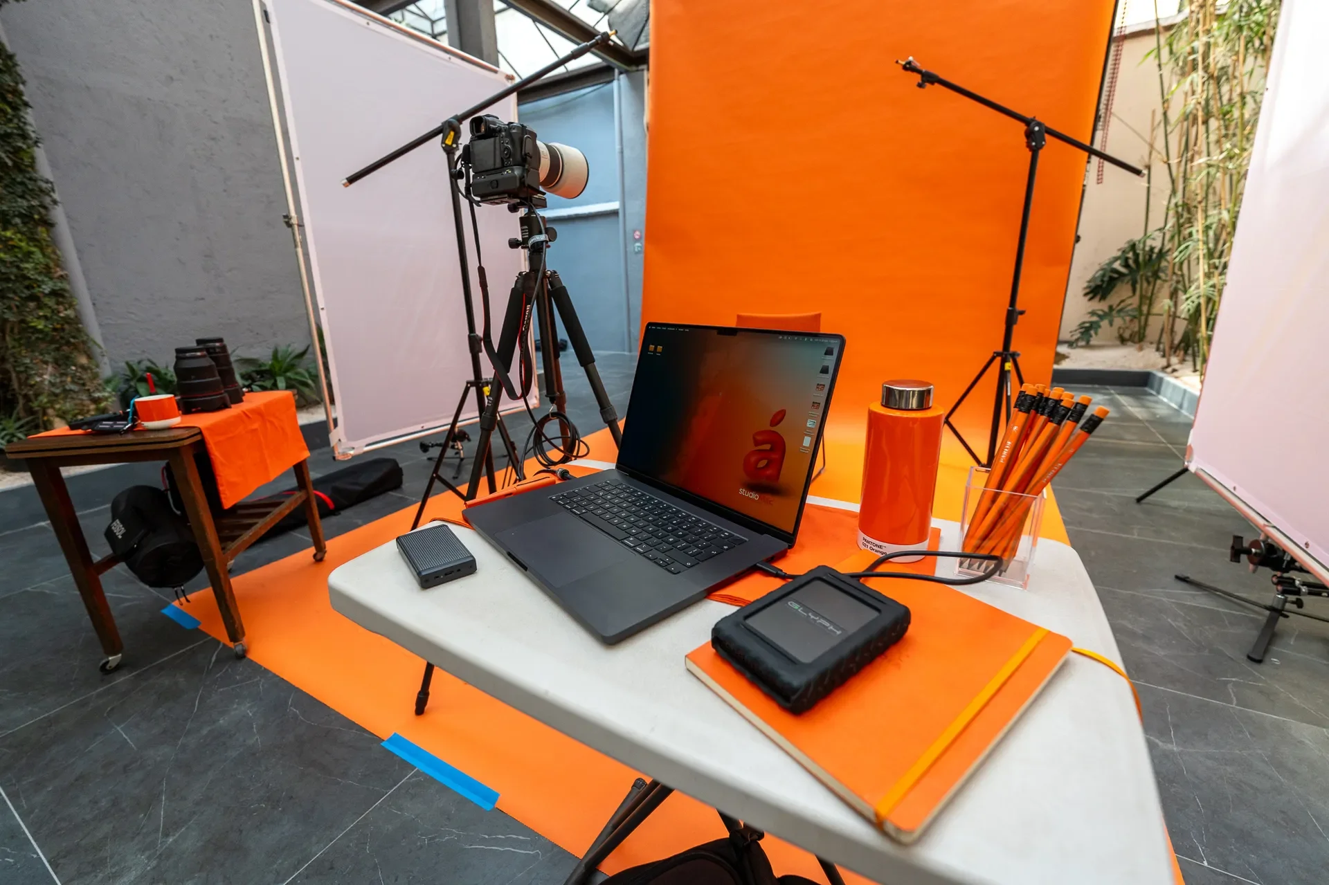 Configuración de estudio fotográfico con fondo naranja, cámara en trípode, micrófono, computadora portátil, y accesorios en una mesa y fondo de estudio.
