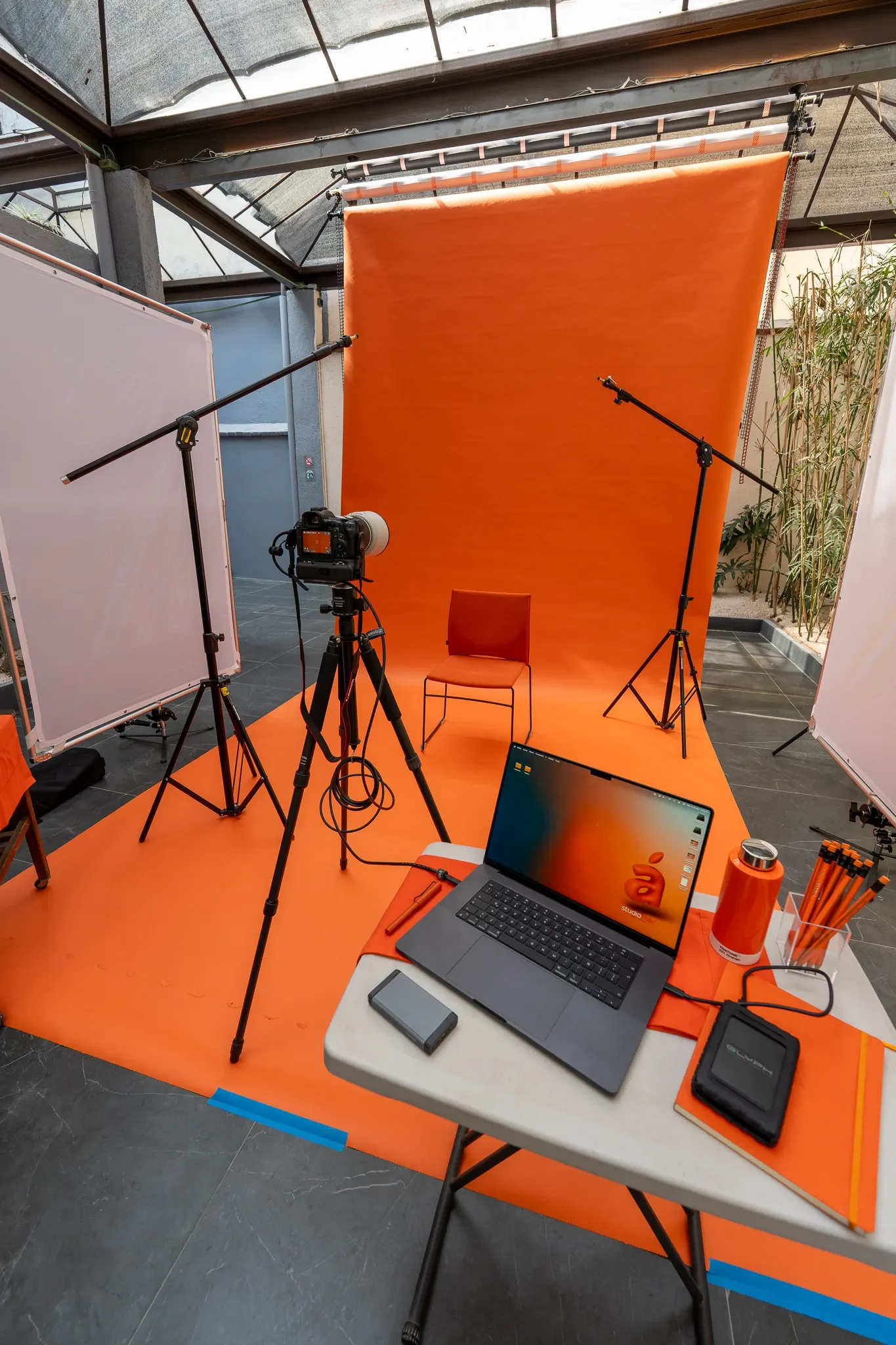 Estudio fotográfico con fondo anaranjado, silla en el centro, equipo de iluminación, cámara en trípode, laptop, y otros accesorios en una mesa.