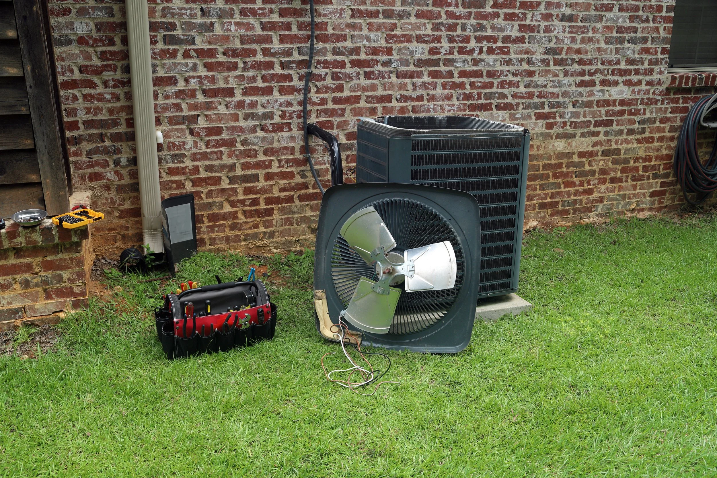 Disassembled outdoor air conditioning unit with fan blade, tools, and wires on grassy yard against brick wall.