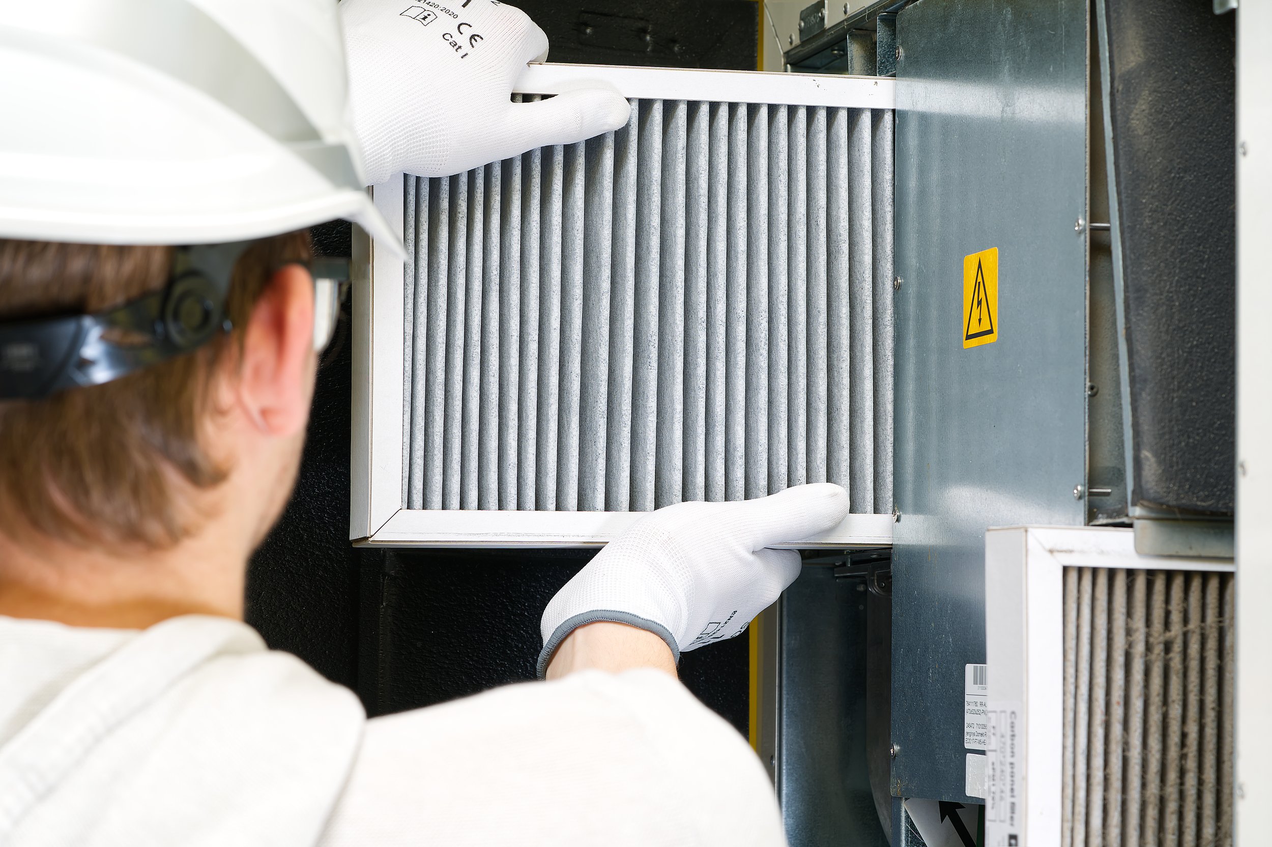 A technician wearing a white hard hat, white gloves, and safety glasses working on a HVAC system, replacing or inspecting a gray filter in an industrial or commercial air conditioning unit.