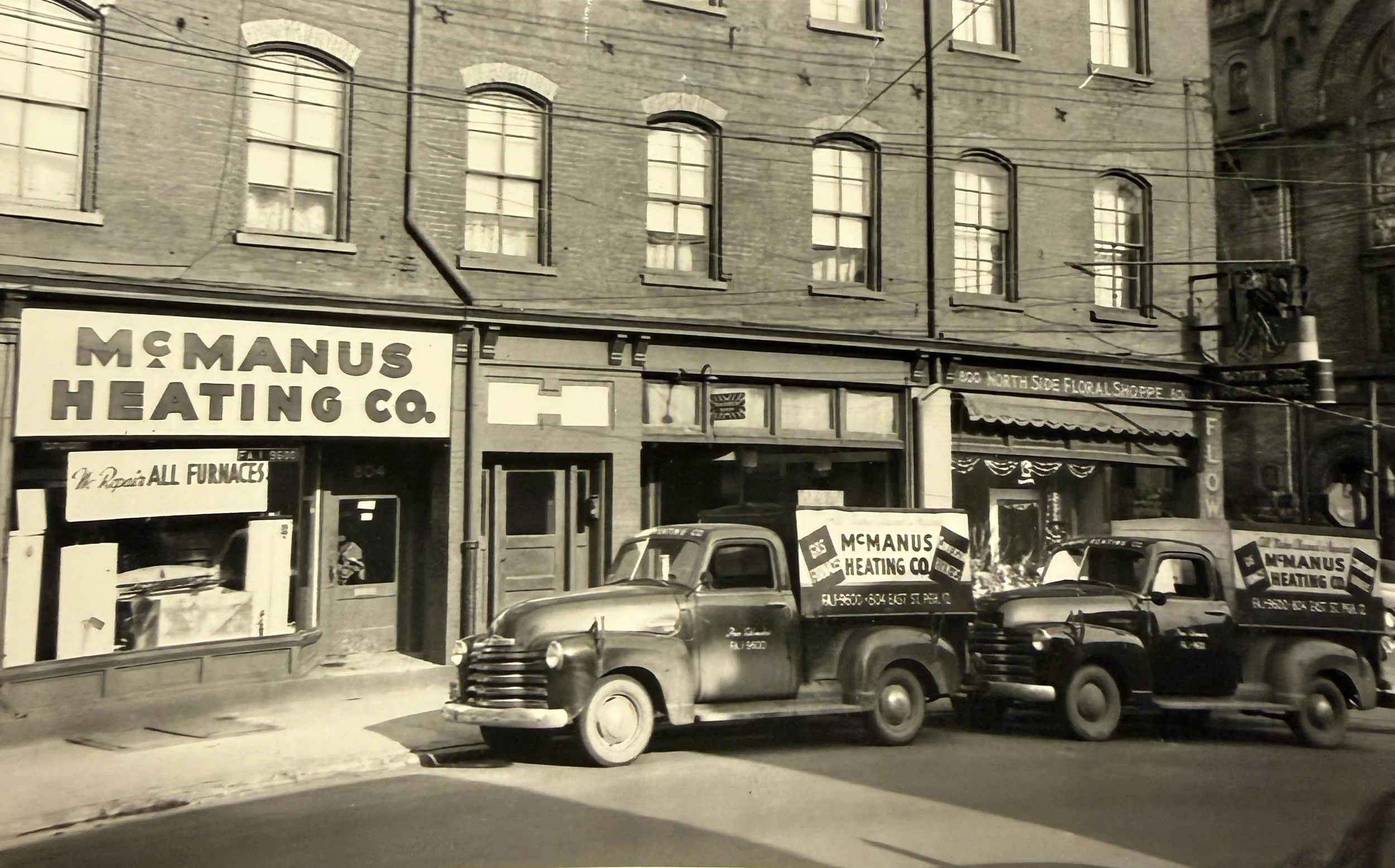 Black and white photo of three vintage trucks parked in front of an old brick building with signs for McManus Heating Co.