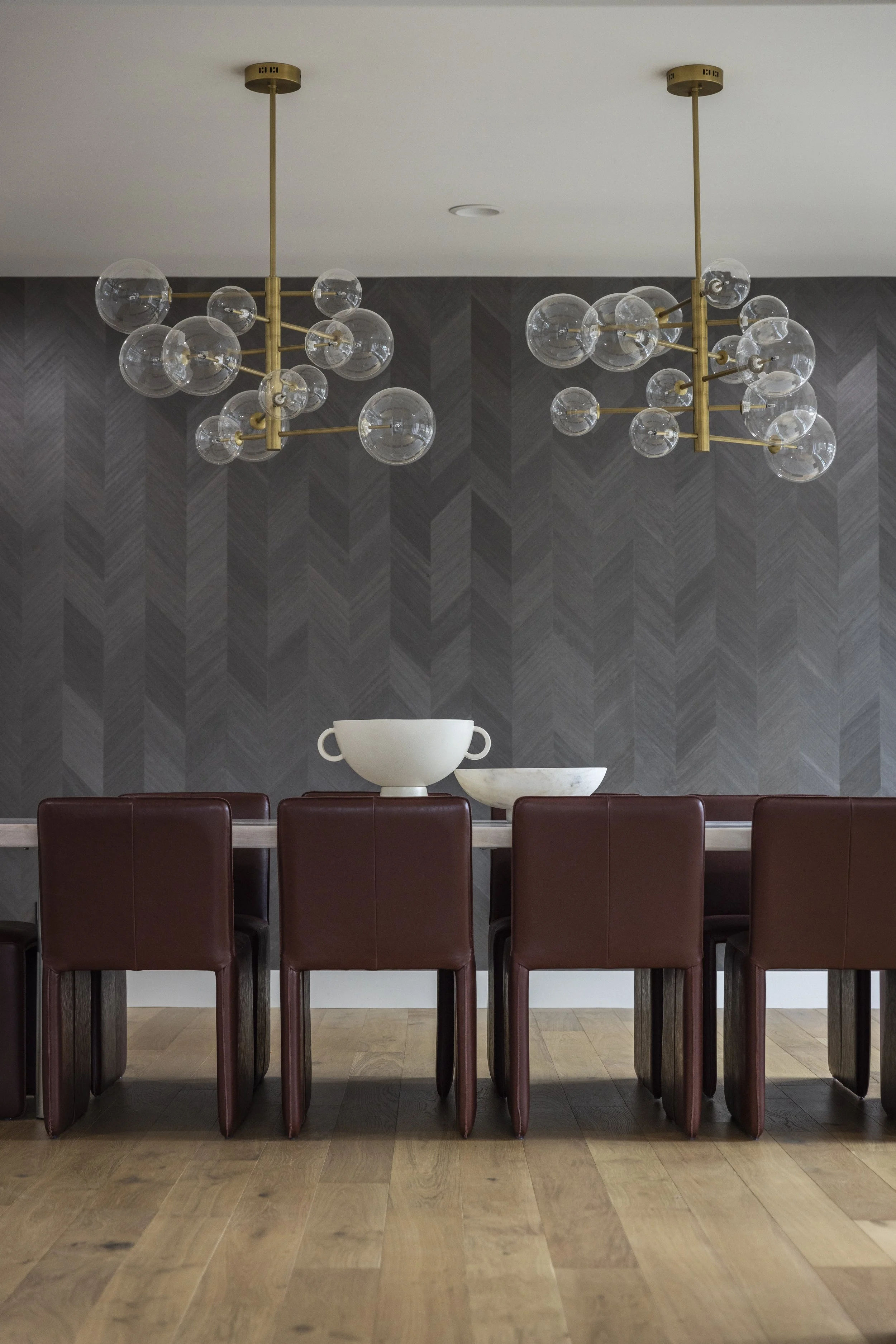 A dining table with six brown leather chairs, three of which are visible. On the table are three white bowls and two plates. Above the table are two modern chandeliers with multiple glass globes and brass fixtures, hanging from the ceiling. The backg