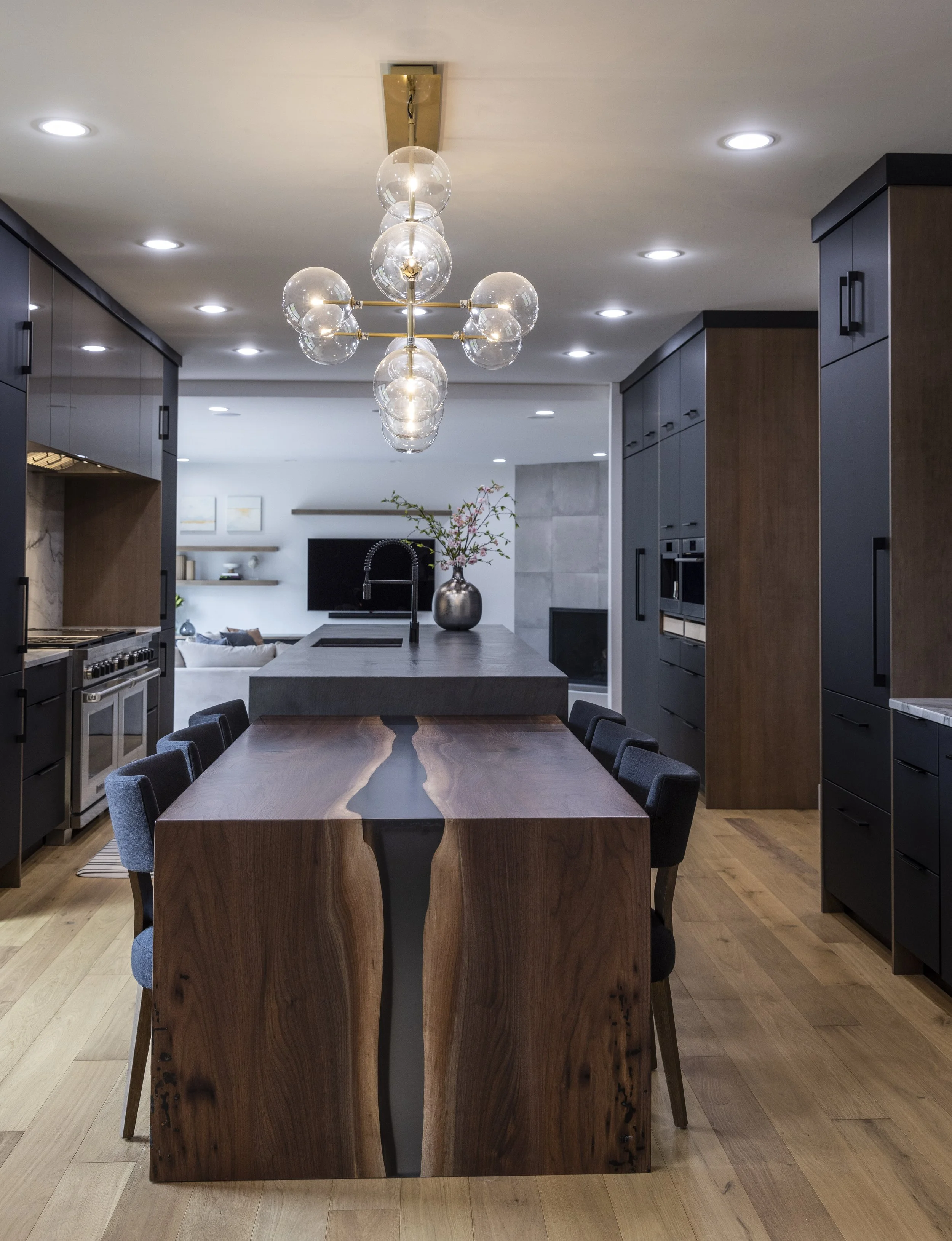 Modern kitchen with wooden dining table, black chairs, black and brown cabinets, and a floral centerpiece. Overhead pendant light with glass globes and recessed ceiling lights.