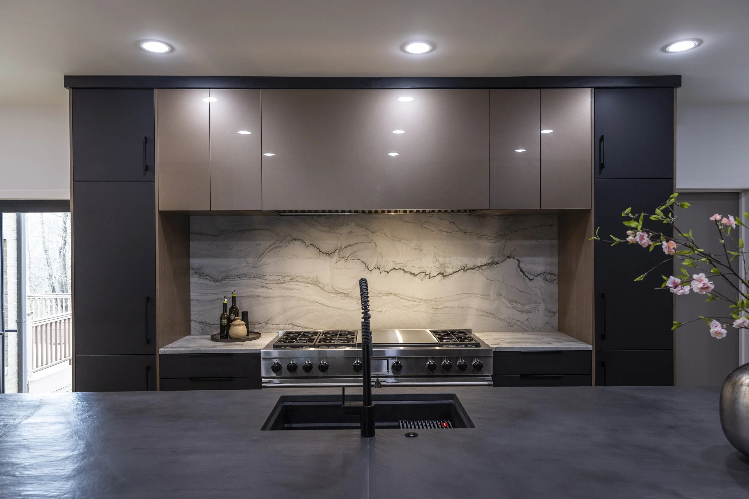 Modern kitchen with black cabinetry, marble backsplash, and stainless steel stove. Overhead recessed lighting, partial view of a window with a deck outside, and a pink flower branch in the foreground.