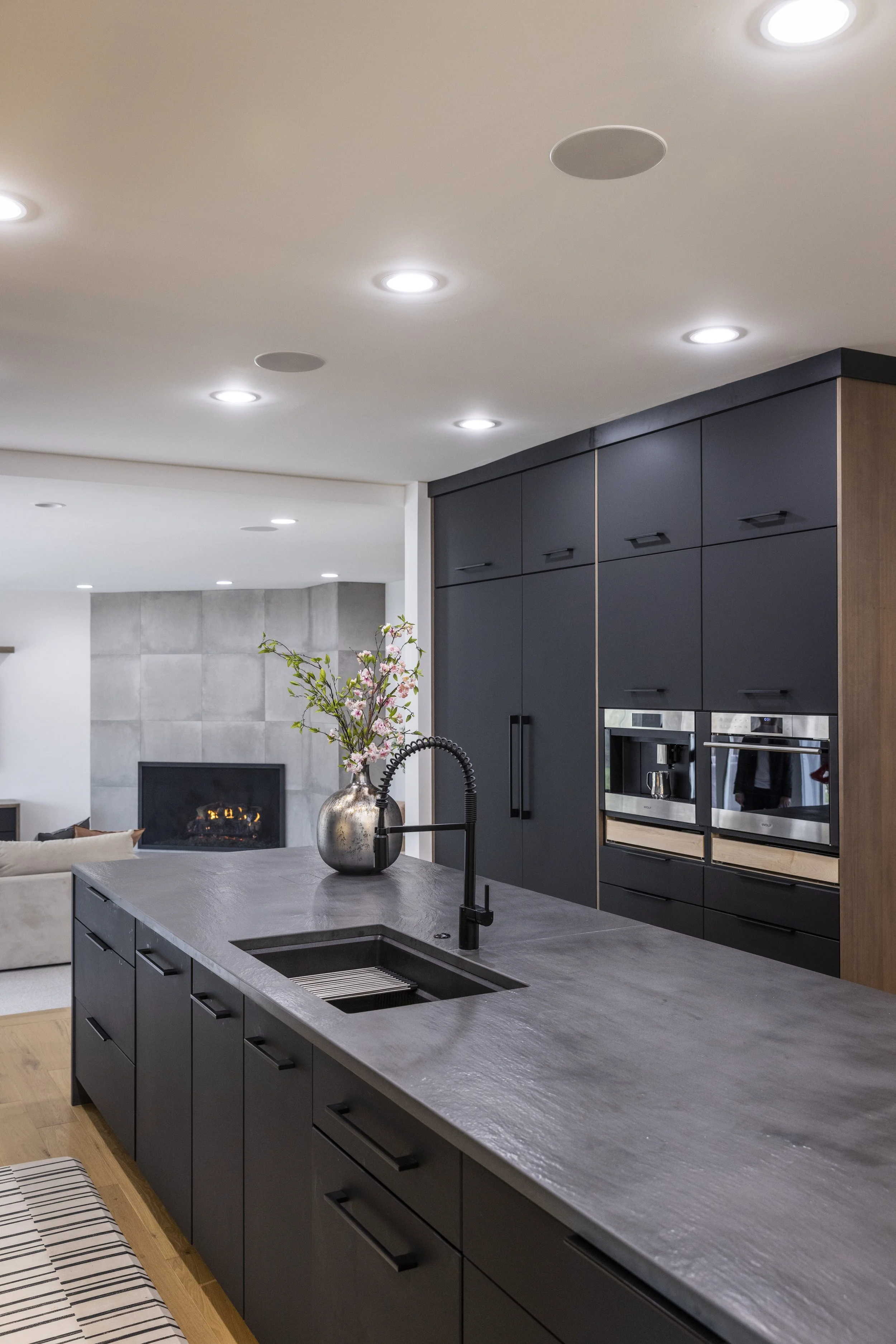 Modern kitchen with black cabinetry, gray stone island with a built-in sink, black faucet, and a decorative vase with pink flowers, in a contemporary open living space with a fireplace and a wall-mounted TV.