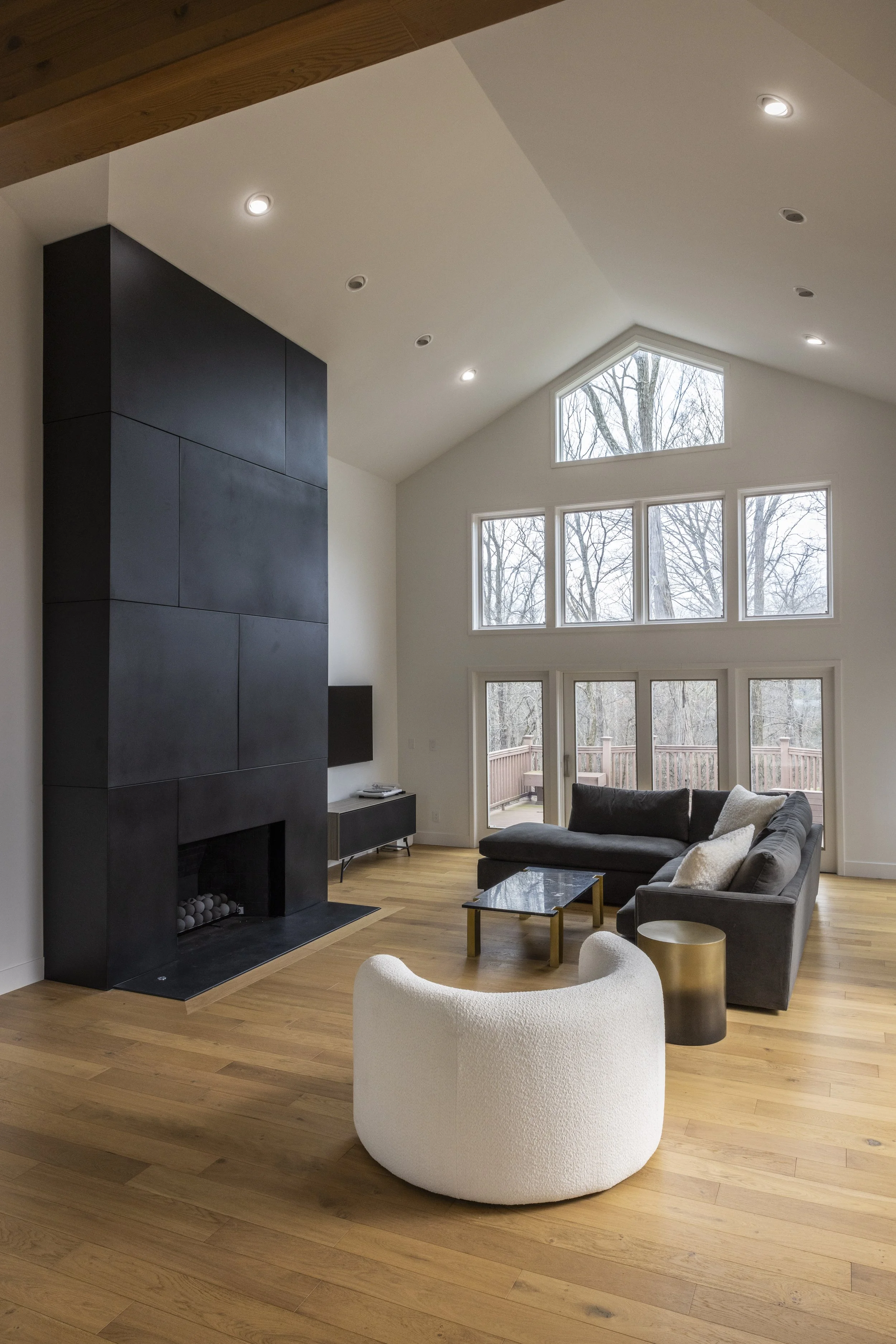 Modern living room with a high ceiling, large windows, a black fireplace, gray sectional sofa, white armchair, and wooden floors.