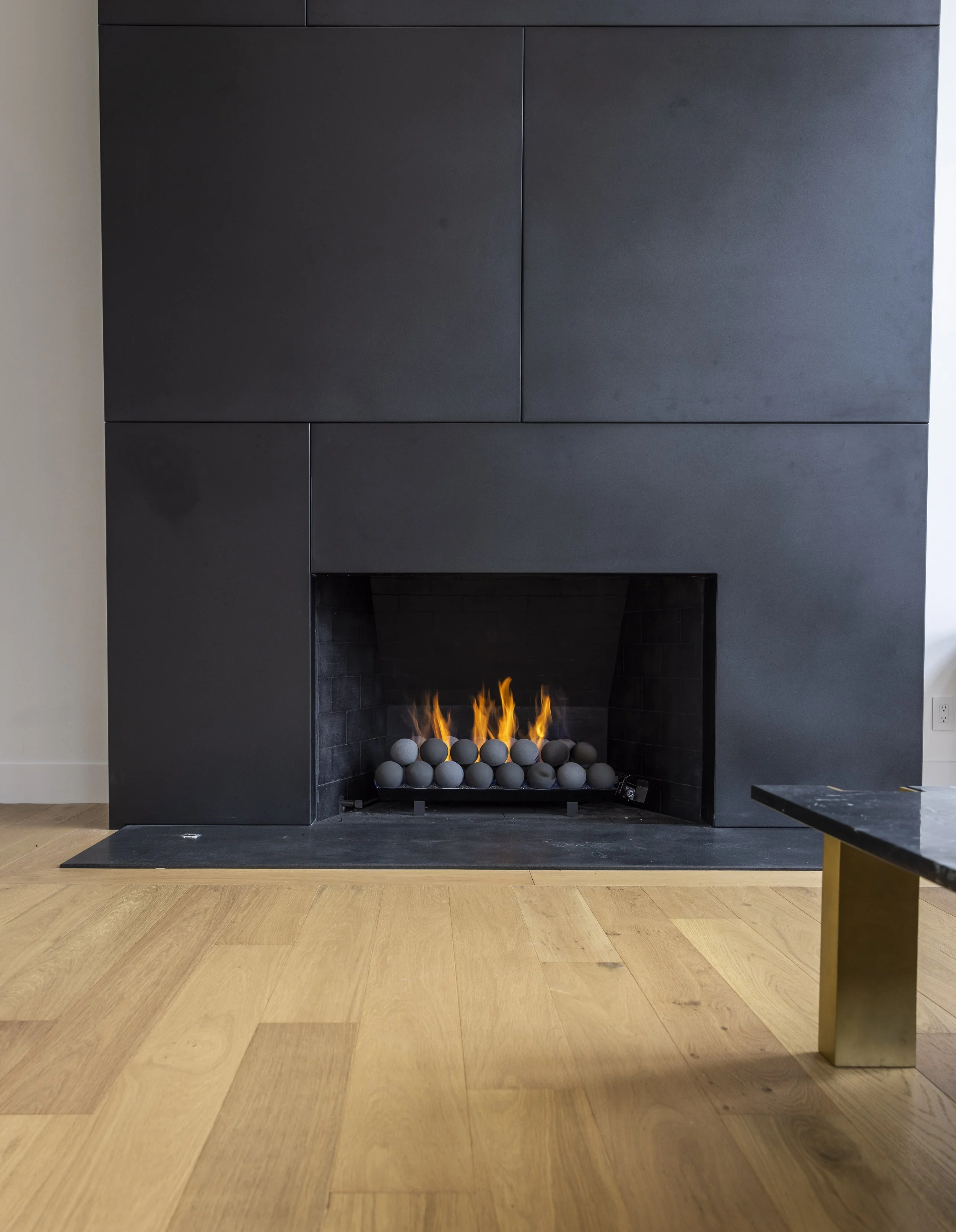 Modern black fireplace with a fire burning inside, surrounded by a black mantel, with a black tiled interior and a light wood floor in front.