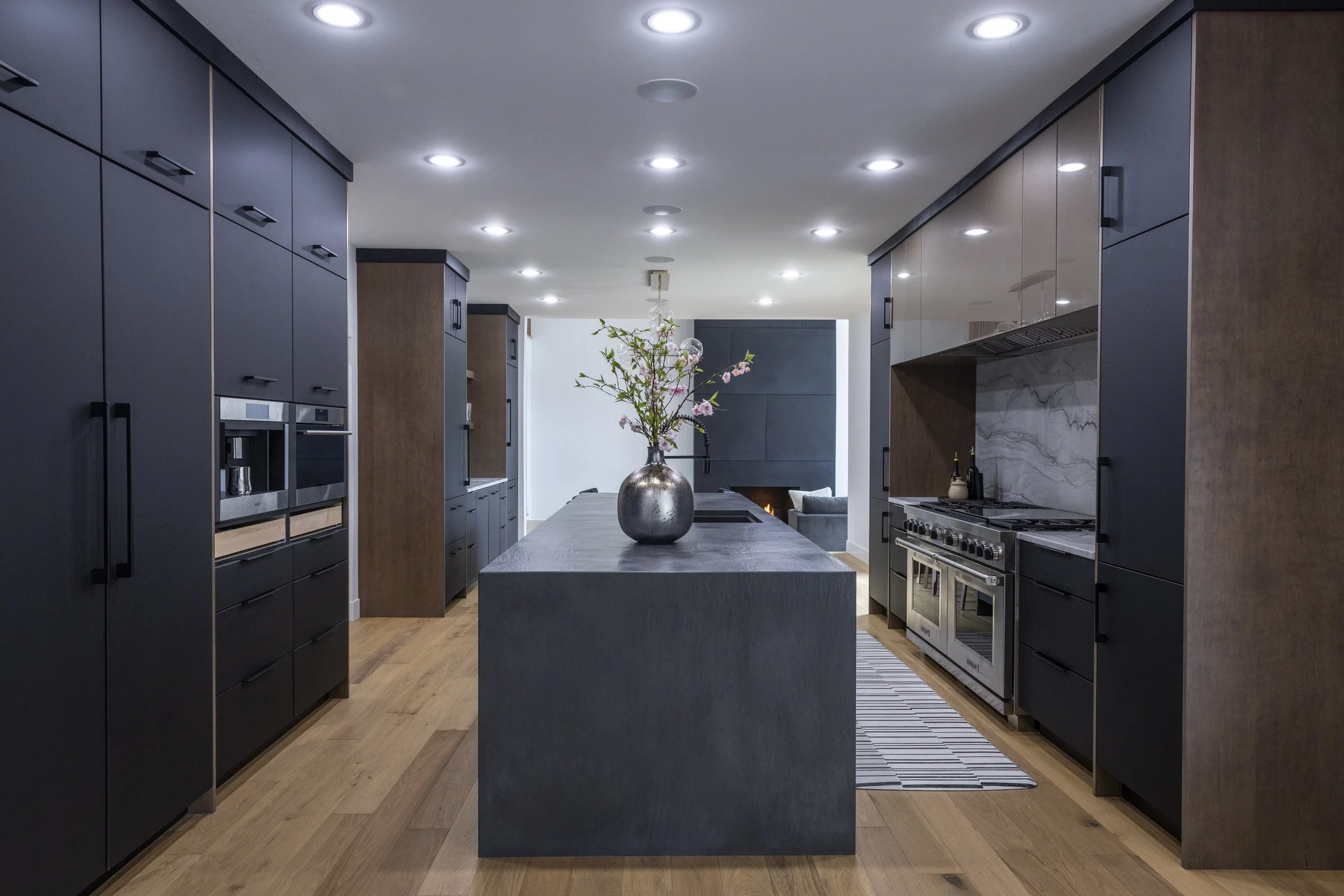 A modern kitchen with dark cabinets, a large gray kitchen island with a vase of pink flowers, stainless steel appliances, wooden flooring, and recessed ceiling lights.