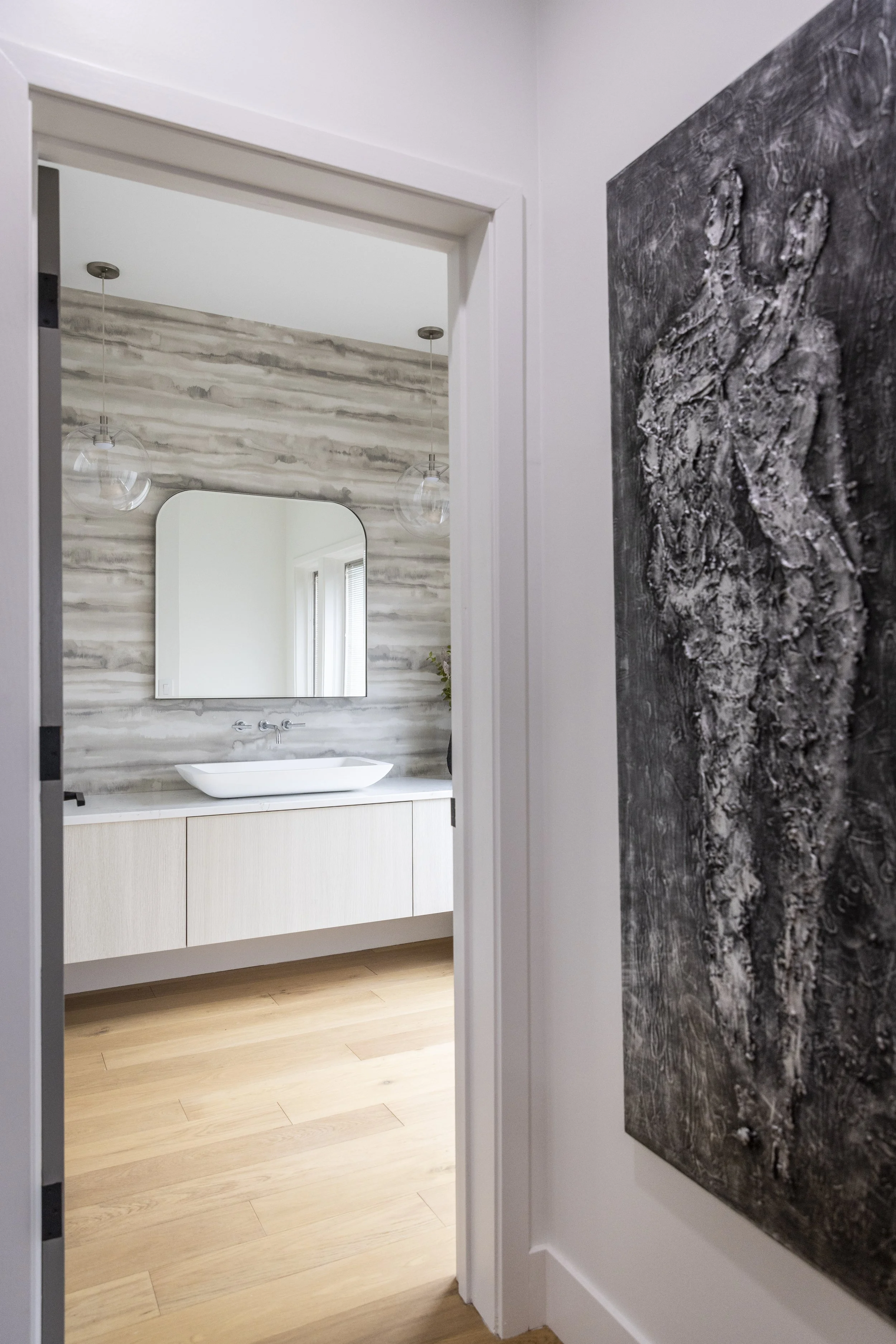 View into a modern bathroom with a floating vanity, a vessel sink, a mirror, striped wall tiles, and two pendant lights, with wood flooring and a marble wall art piece on the right.