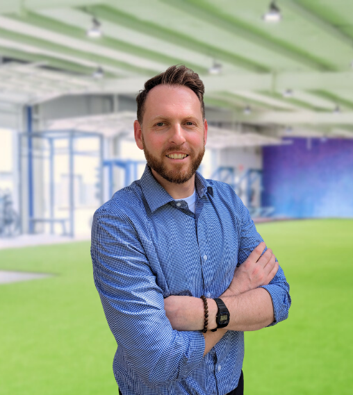 A smiling man with a beard and short hair, wearing a blue patterned button-up shirt, standing outdoors with a modern building and green lawn in the background.