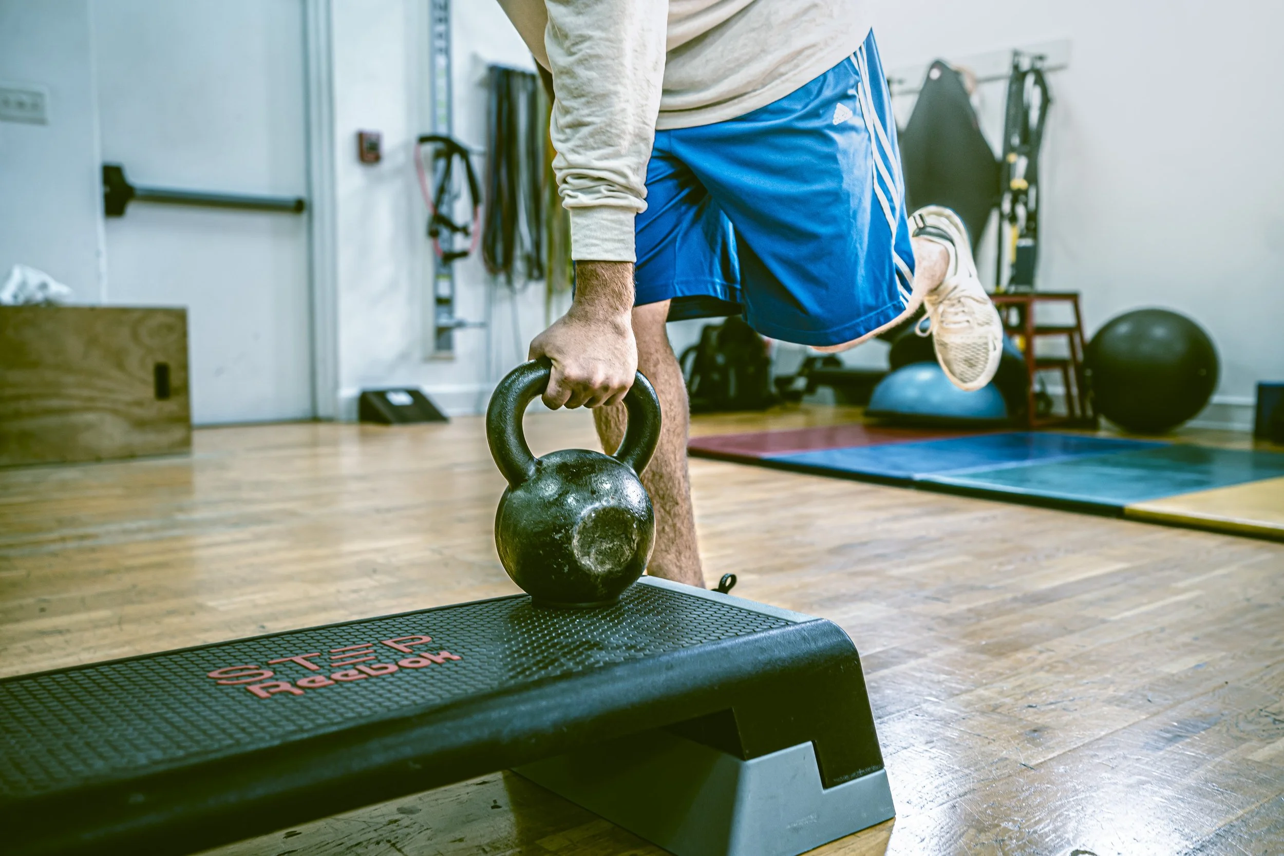 Man performing a step-up exercise with a kettlebell in a gym, wearing blue shorts and a long-sleeve shirt.