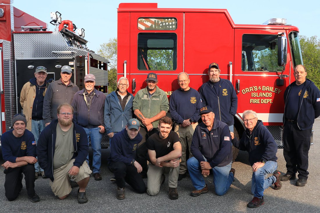 Group of firefighters and volunteers posing in front of a red fire truck with 'Orr's & Bailey Islands Fire Dept' written on it, outdoors on a clear day.