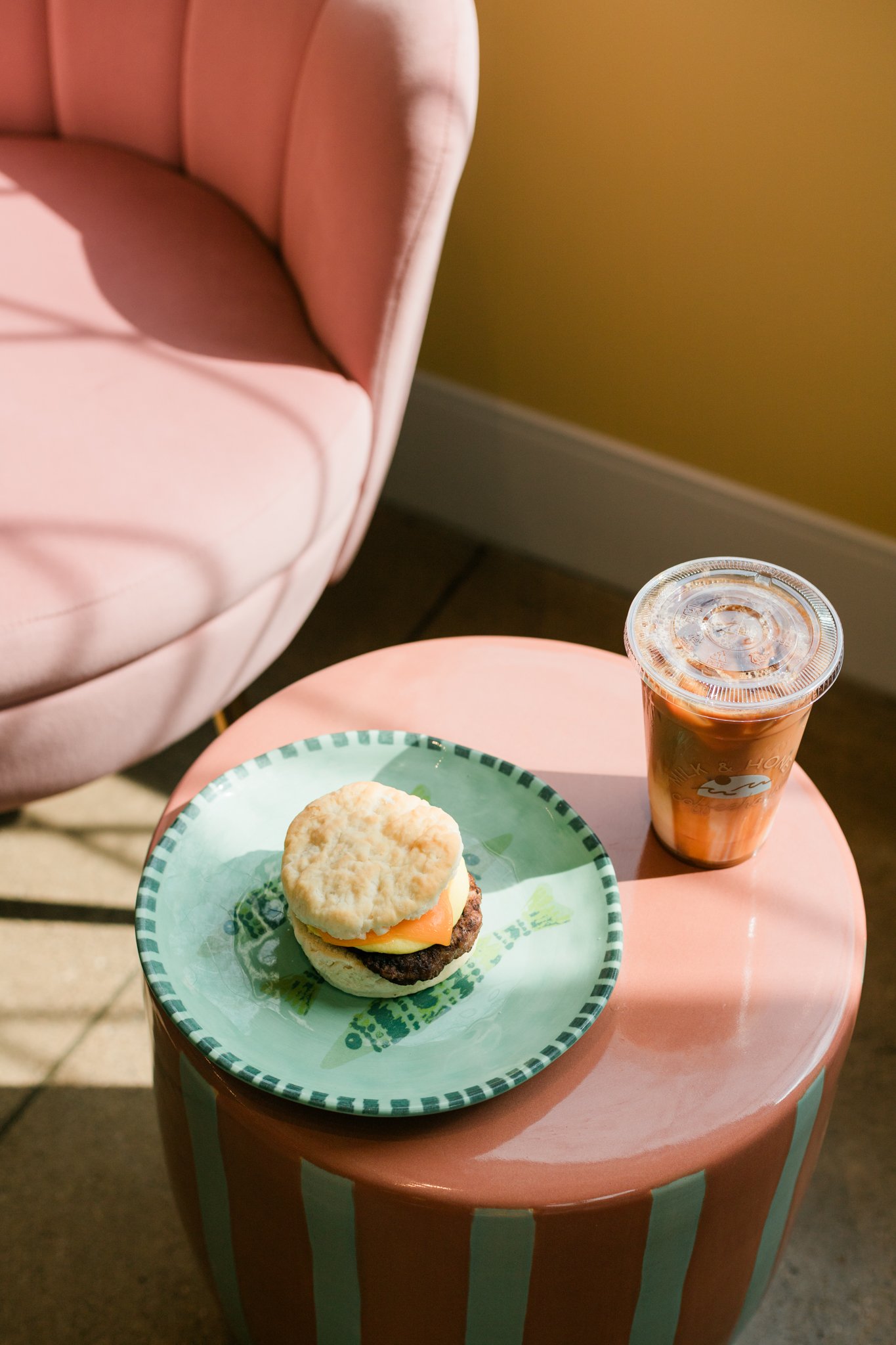 A small pink table with a plate holding a cheeseburger and a plastic cup of iced coffee or soda on it, beside a pink upholstered chair.