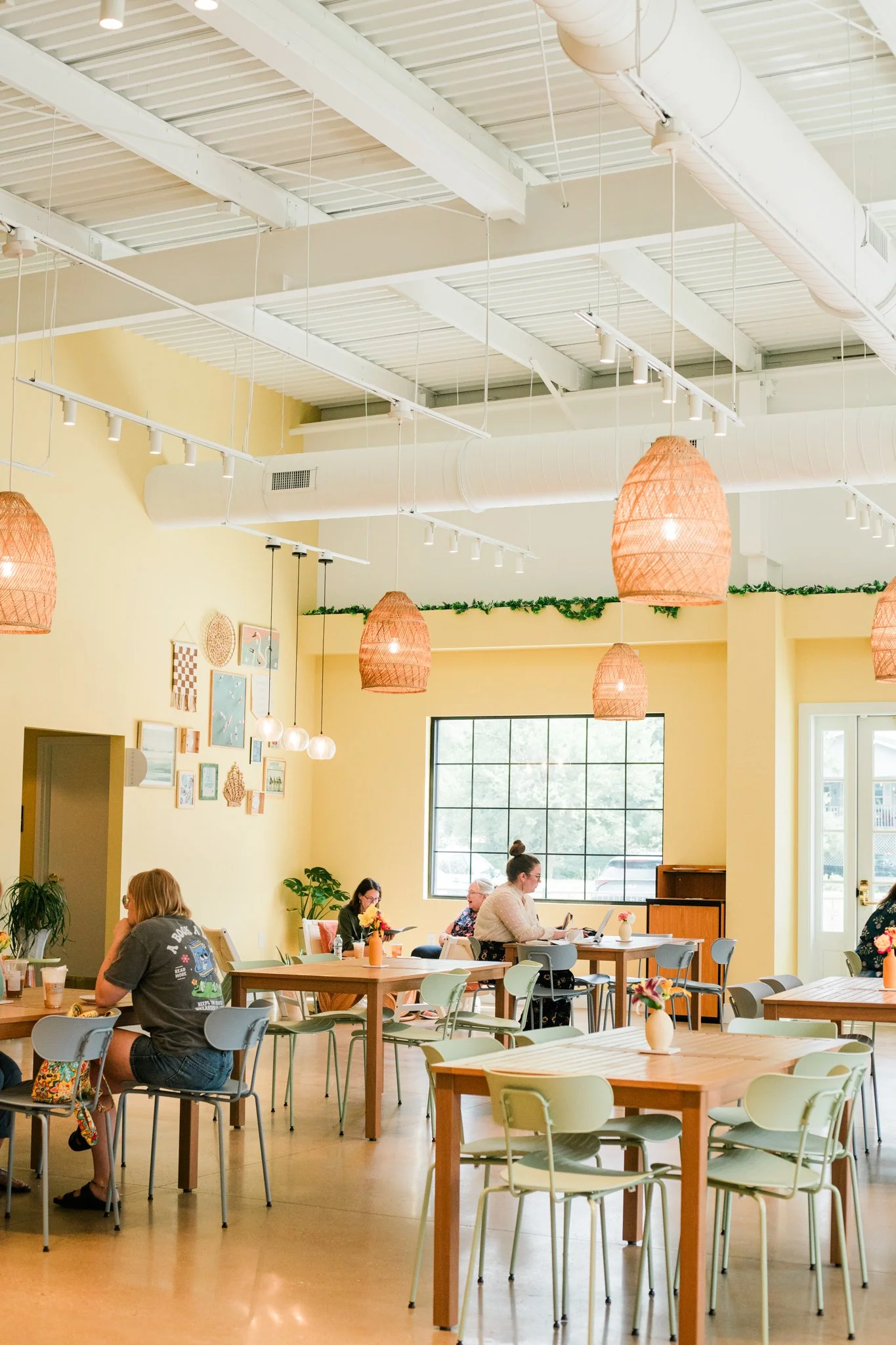 Interior of a bright, airy cafe with yellow walls, hanging wicker light fixtures, large window, and several people seated at tables.