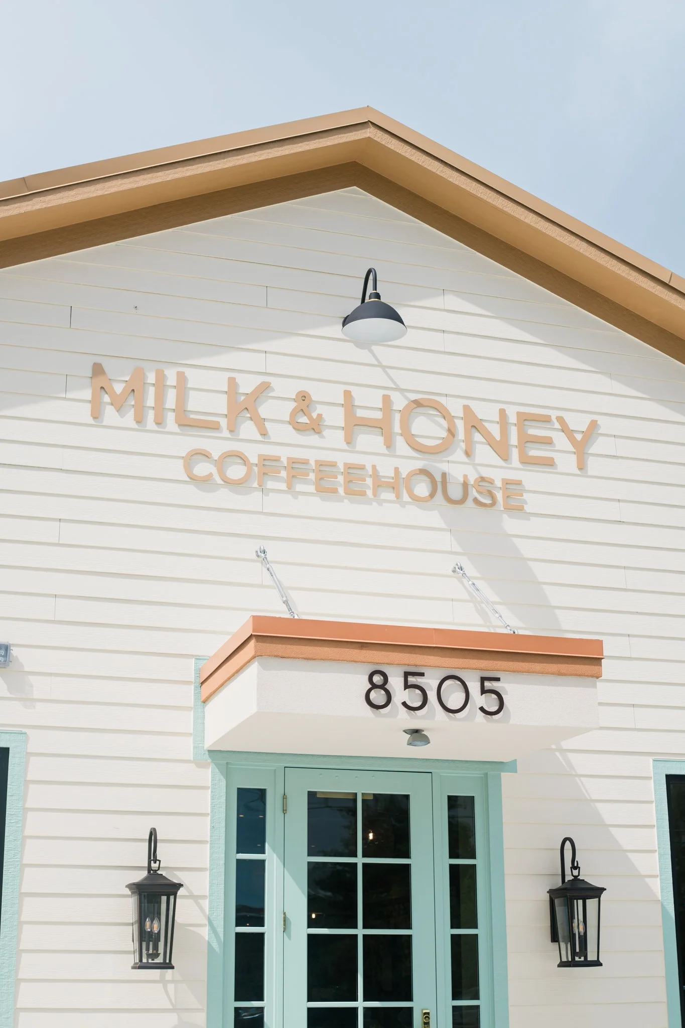 Exterior of a building with a sign that reads "Milk & Honey Coffeehouse" and the address 8505 above the door, which is painted in a light teal color with glass panes. The building has white siding and is decorated with black lantern-style wall lights.