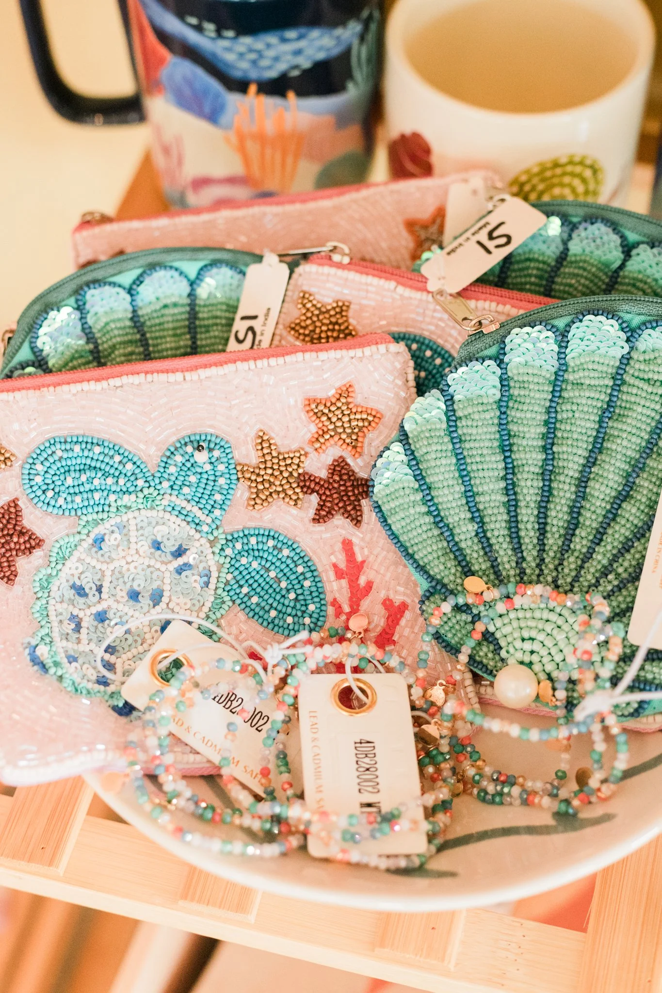 Colorful beaded jewelry, including bracelets, displayed in small pouches with ocean and shell designs, placed in a bowl. In the background, there are two mugs.