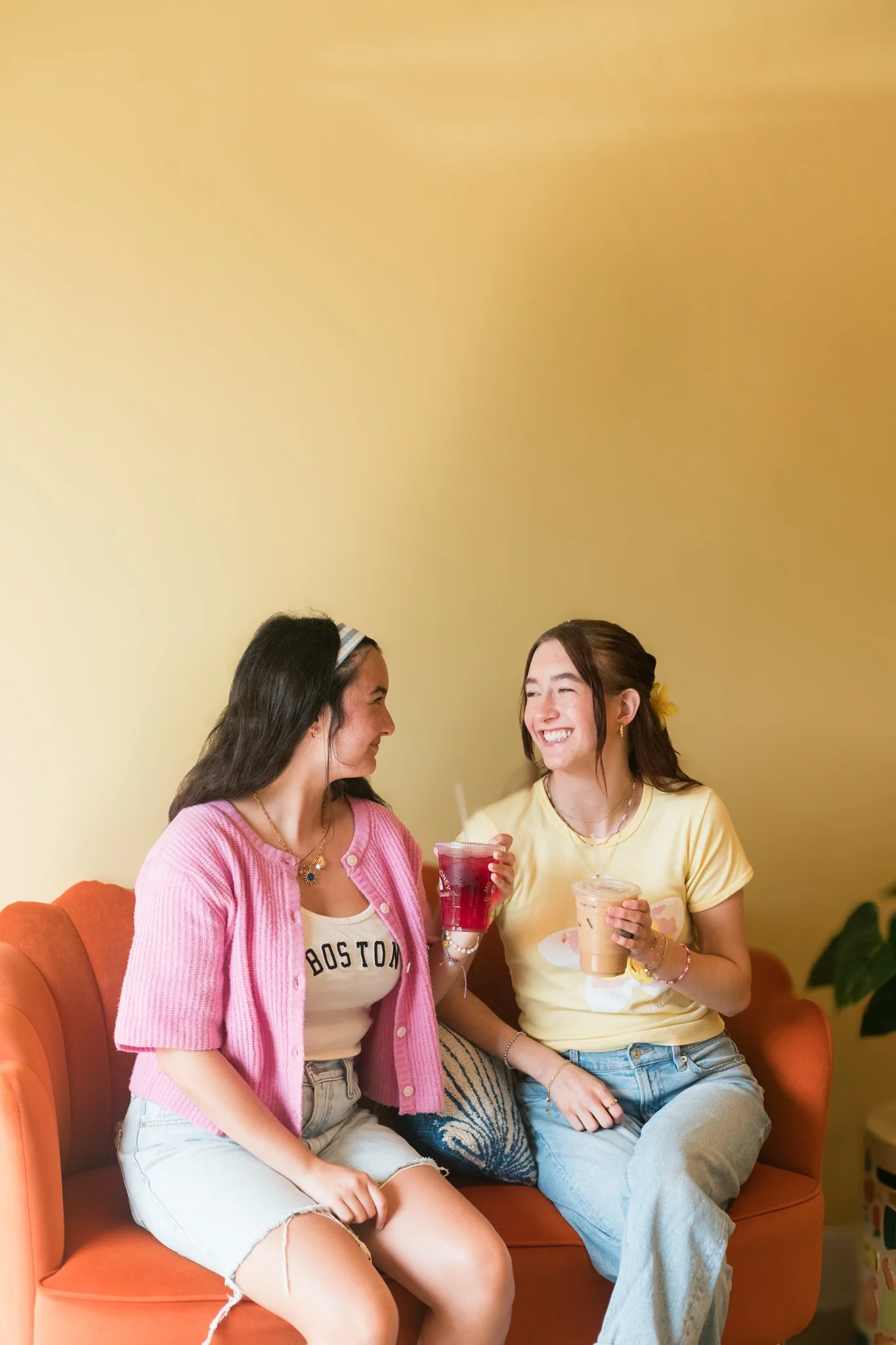 Two women sitting on an orange sofa, smiling and drinking beverages, having a conversation in a cozy indoor setting.