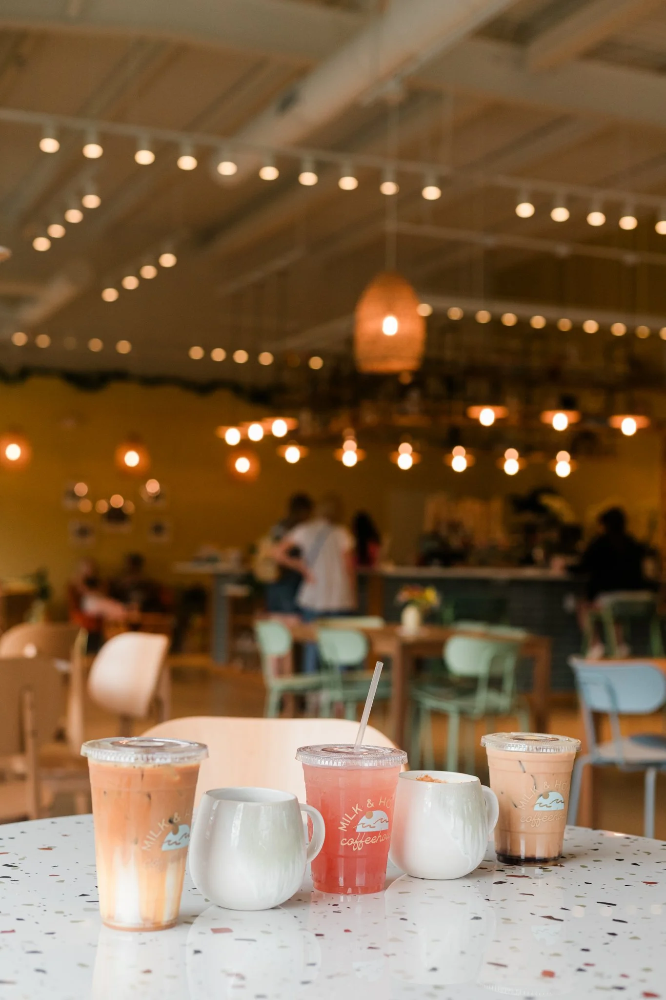 Three iced drinks with straws and two white ceramic mugs on a speckled white table in a cozy cafe, with blurred warm lighting and people in the background.