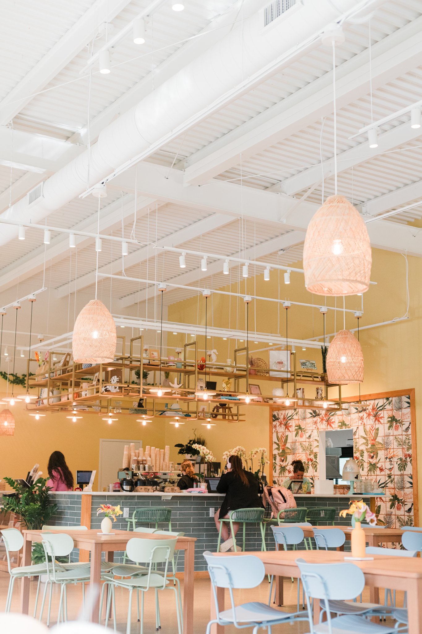 Bright and cheerful cafe interior with pendant lights, a barista counter, and pastel-colored chairs and tables, decorated with plants and floral artwork.