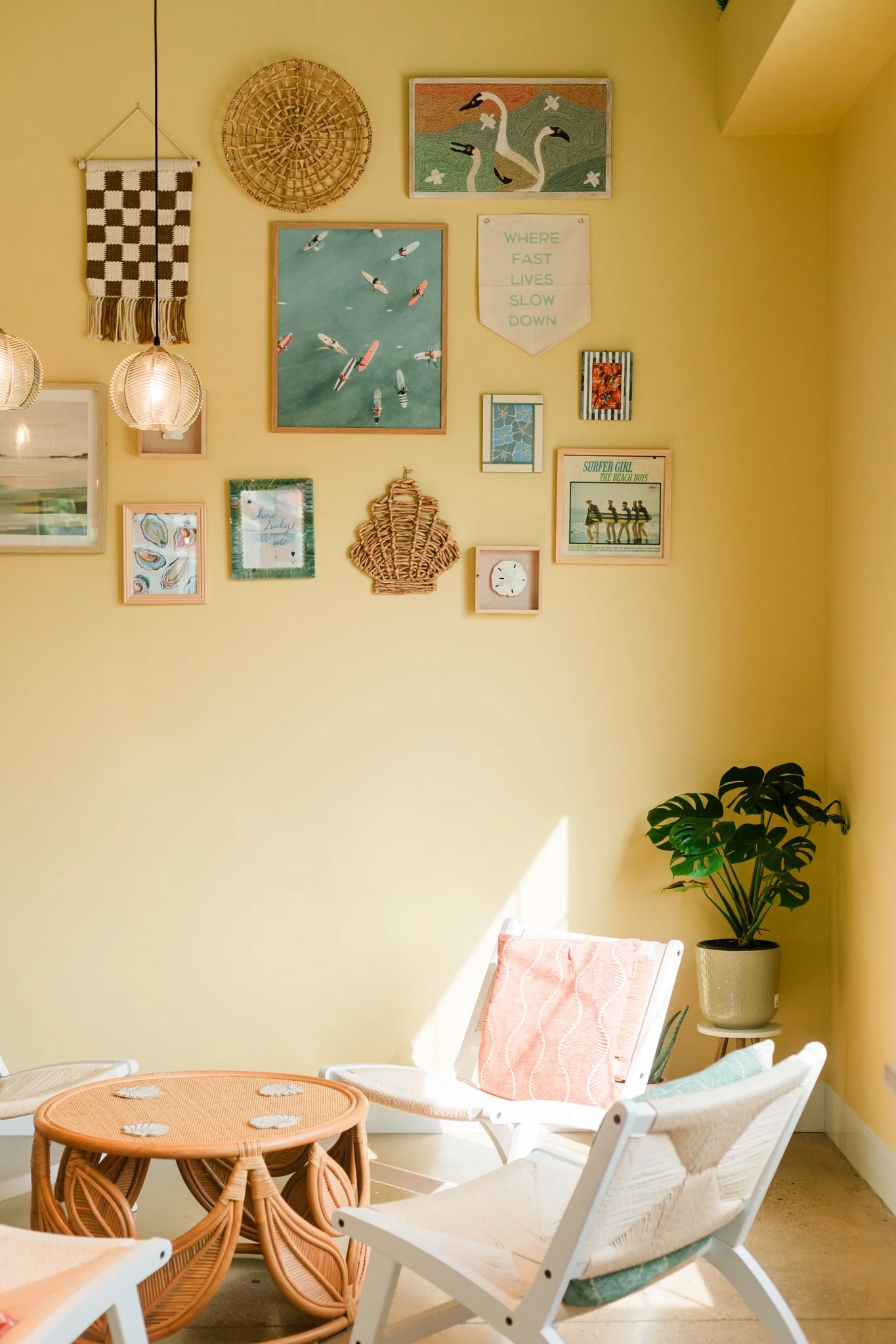 Sunlit corner with yellow walls, decorated with framed art and woven wall hangings, white chairs with pastel cushions, a round wicker coffee table, potted Monstera plant.