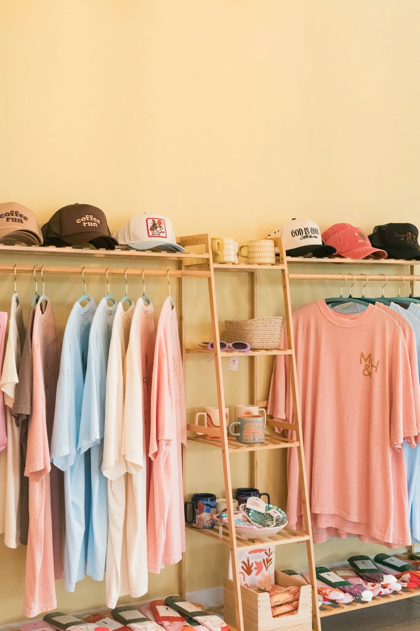 Display of pastel-colored T-shirts, hats, mugs, and accessories on a clothing store shelf with a light yellow wall.