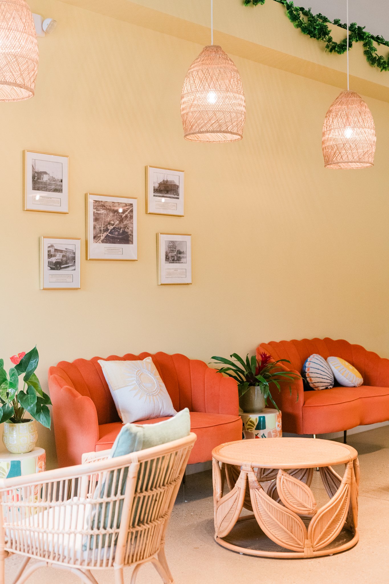 A cozy waiting area with two coral-colored sofas, decorative pillows, potted plants, a rattan coffee table, wall art, and hanging woven light fixtures.