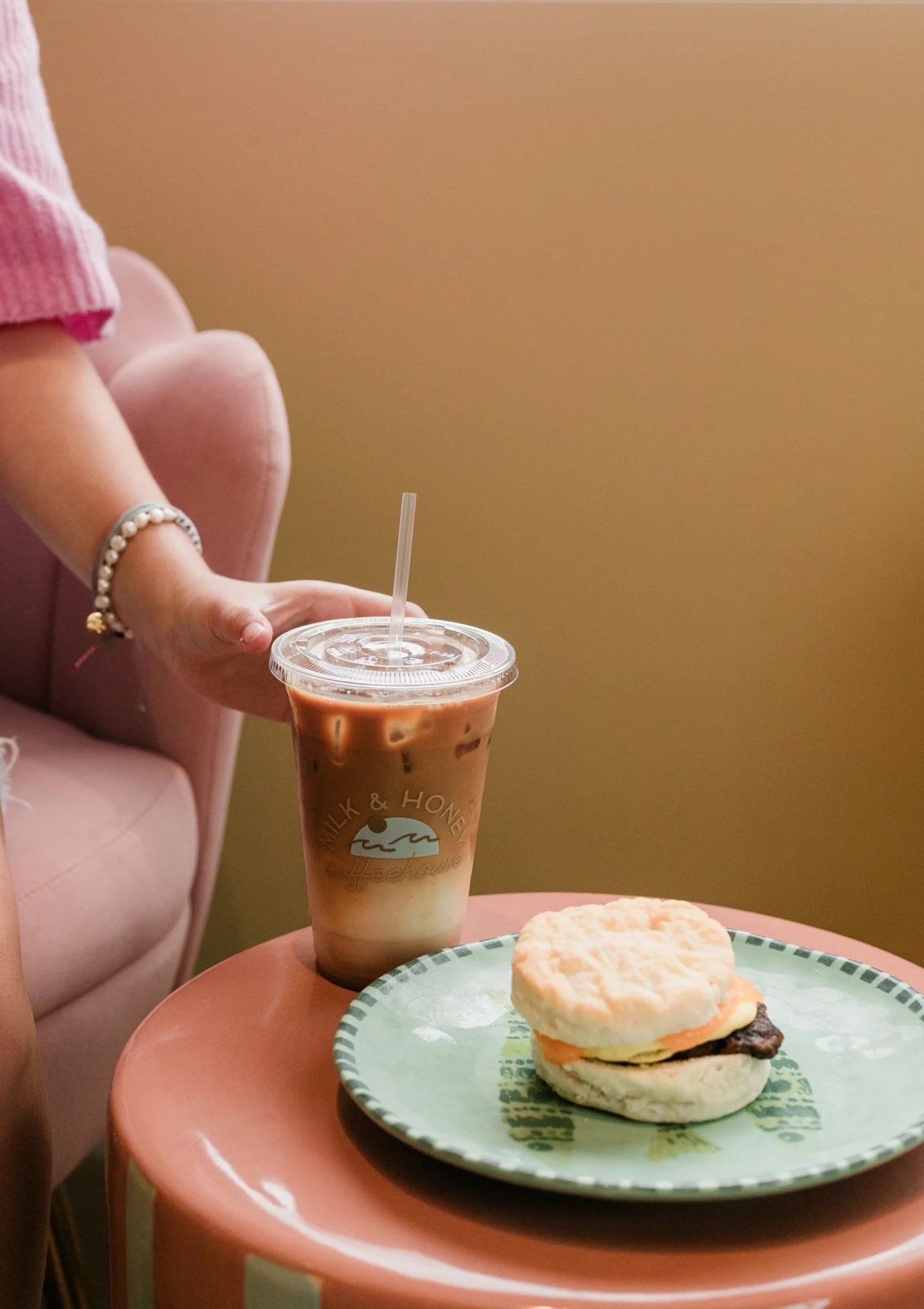 A hand with a pearl bracelet reaching for a cup of iced coffee on a pink side table, with a sandwich on a green plate nearby.