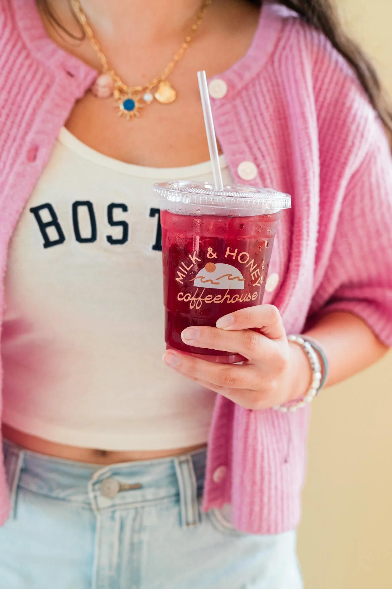 Person holding a plastic cup with iced red drink from Milk & Honey Coffeehouse, wearing a pink cardigan, white tank top with 'BOSTON' written on it, and jeans.
