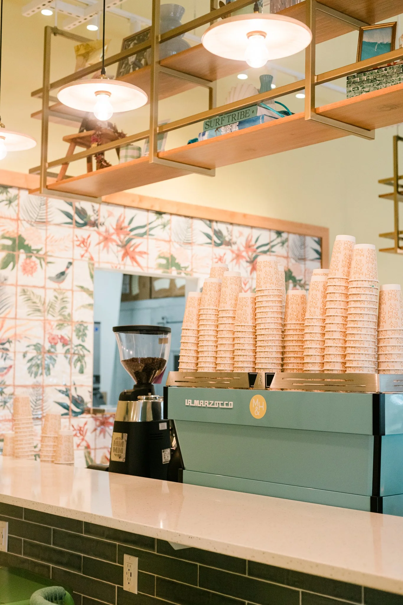 A coffee shop counter with a green espresso machine, stacks of pink paper coffee cups, a coffee grinder with coffee beans, and wooden shelving with decorative items and books, in a bright, modern space with palm leaf patterned tiles and pendant lighting.