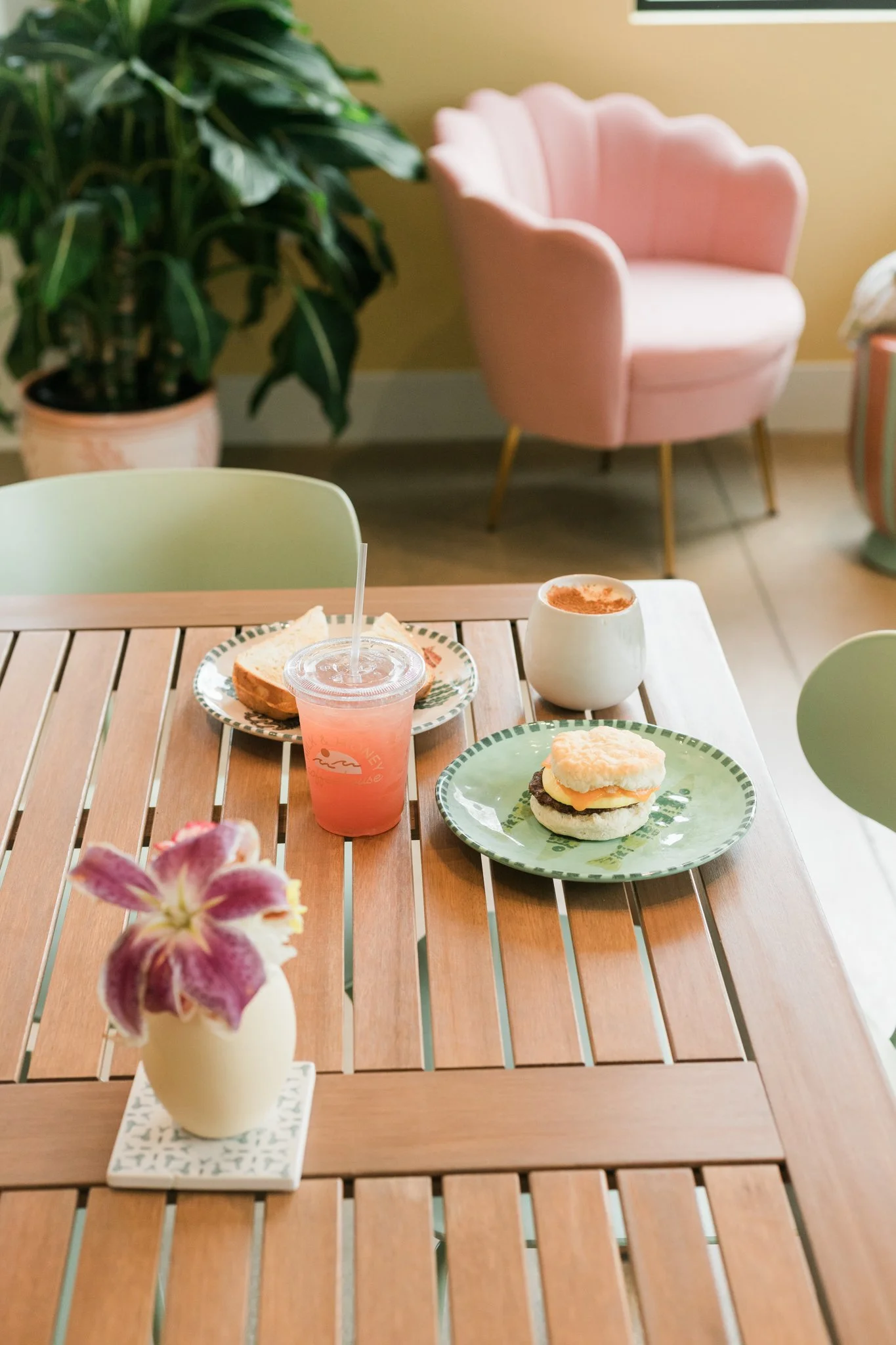 A decorated wooden table with a pink drink, a cupcake, a sandwich, and a plant with a pink flower in a white vase, set in a cozy room with pastel-colored chairs and plants.