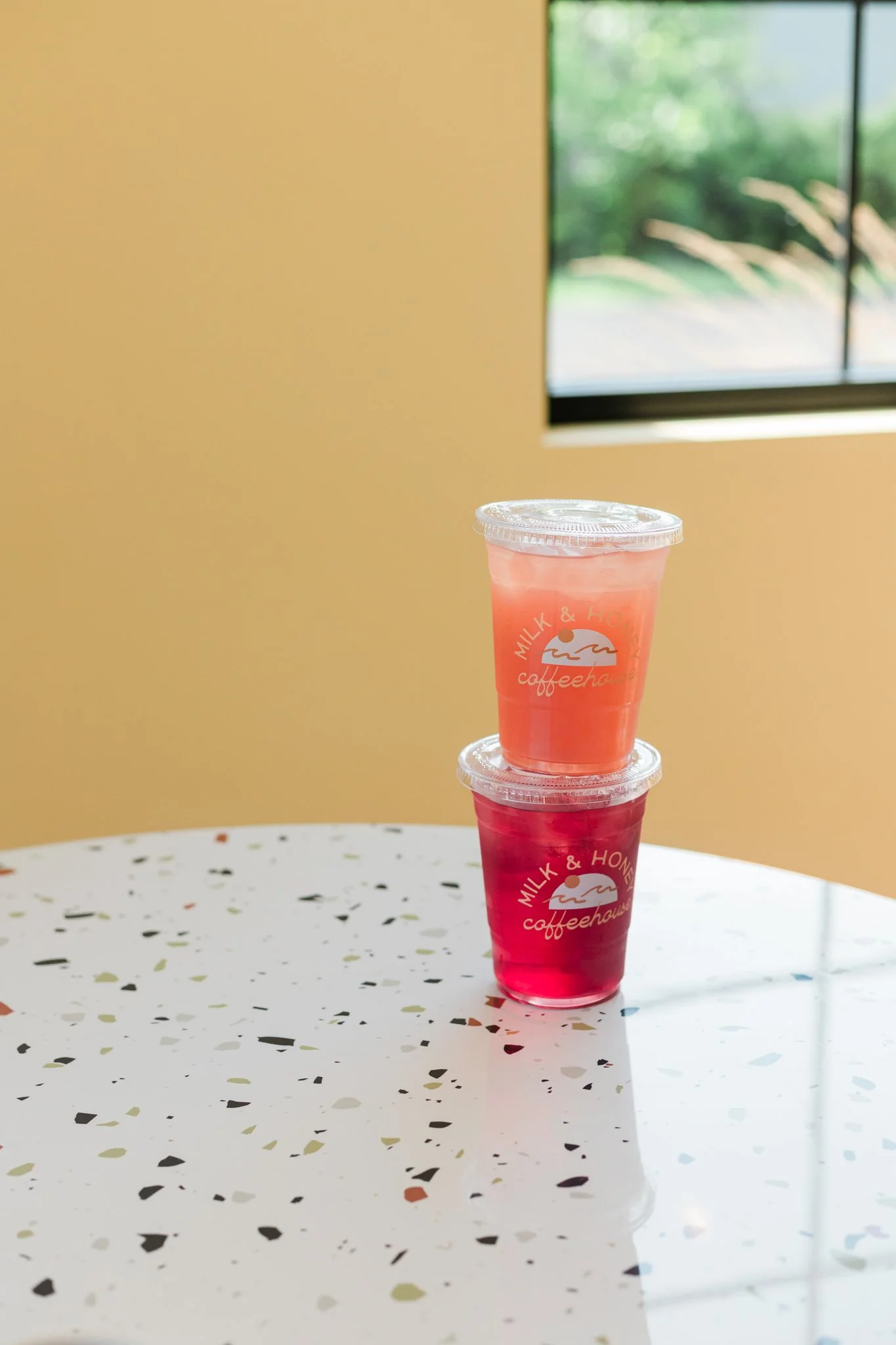 Two cups of pink drinks stacked on a terrazzo table inside a cafe with a window showing green trees outside.