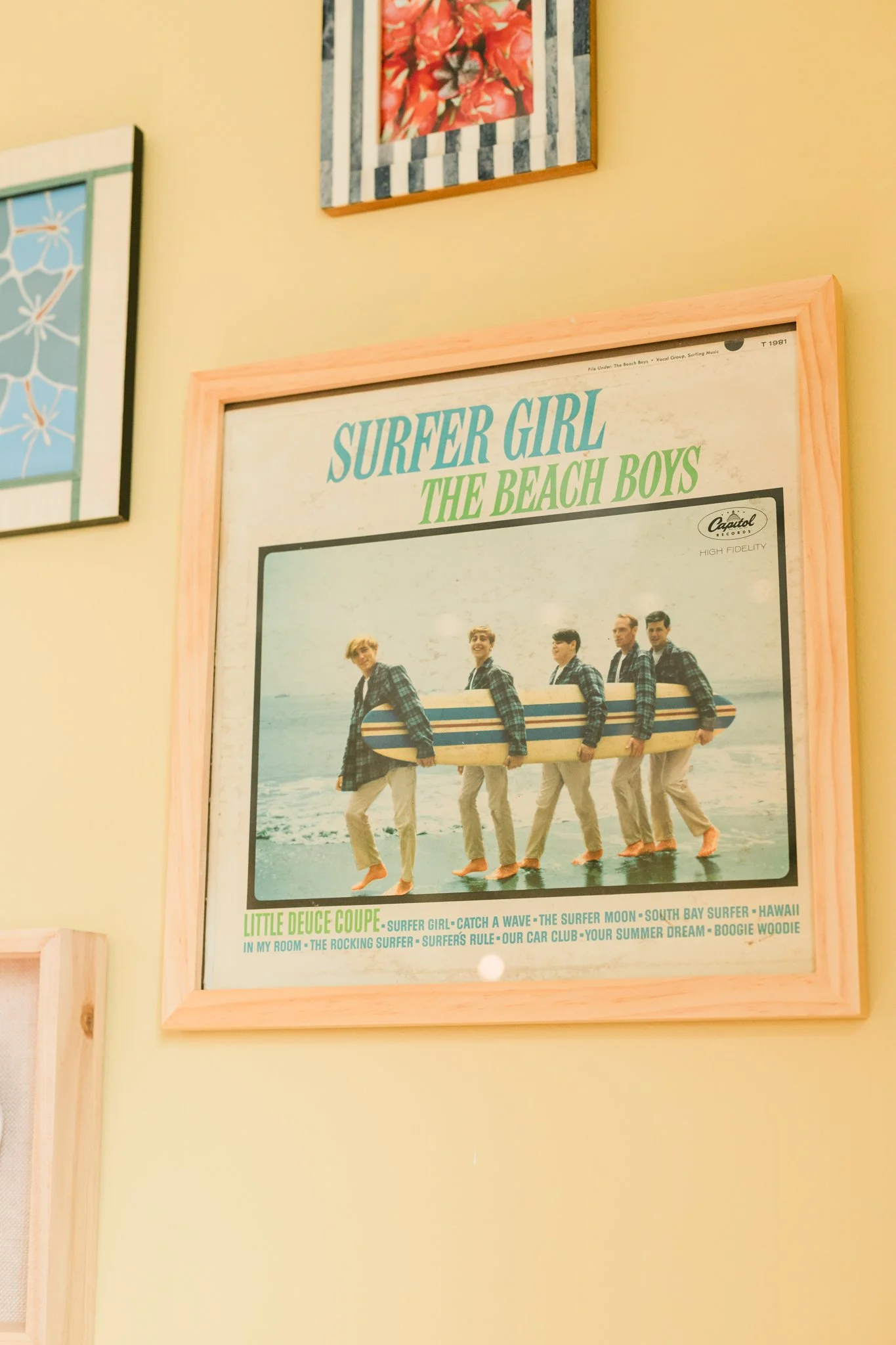 A framed vintage poster titled 'Surfer Girl' by The Beach Boys, showing five young men at the beach carrying a surfboard, with a yellow wall and other framed artwork visible around it.