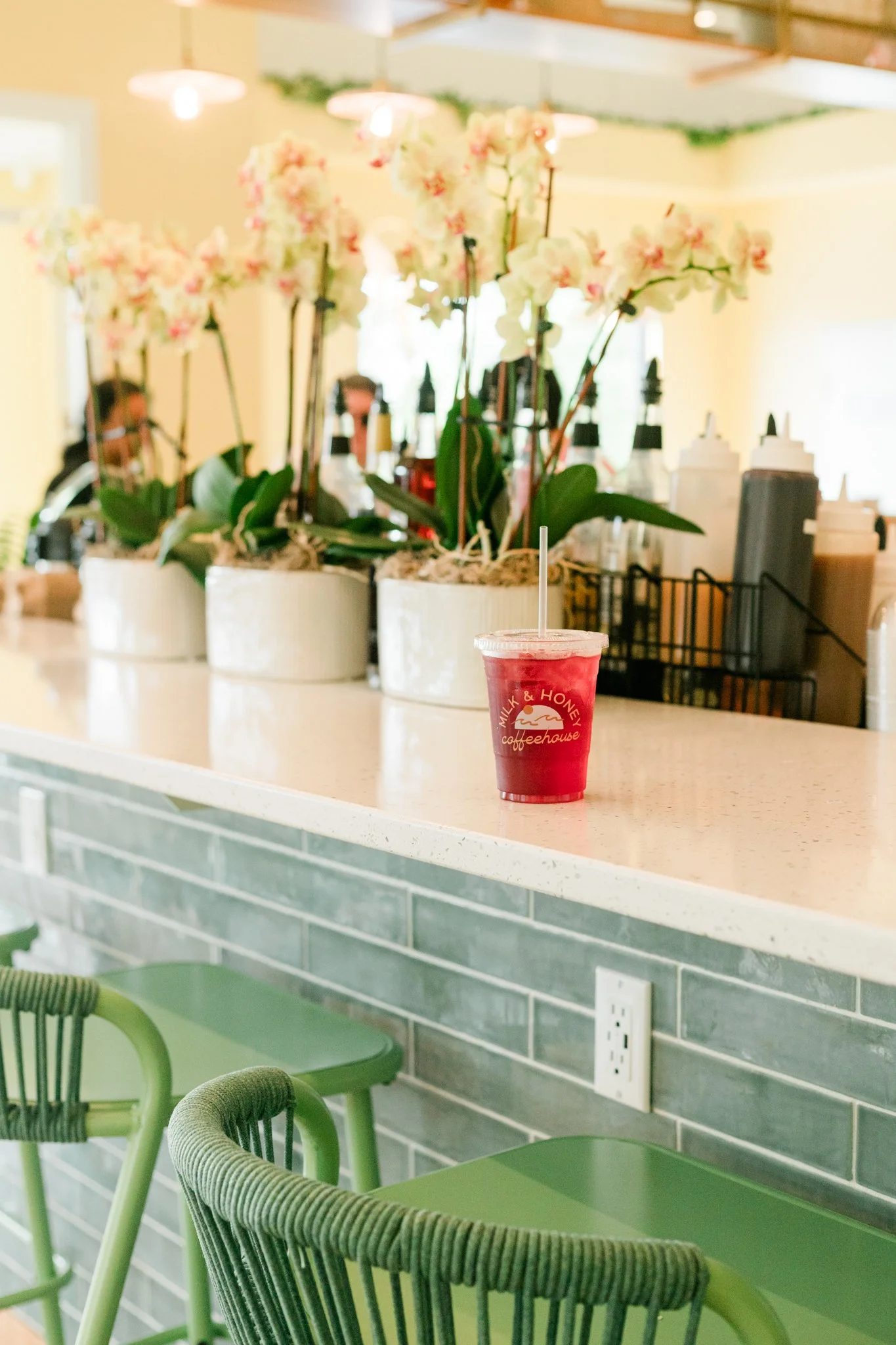 A red iced beverage in a clear plastic cup with a straw is placed on a white marble countertop in a cafe. Behind the cup are potted pink orchids and assorted condiment bottles, with a green brick wall below the counter and green chairs in the foreground.