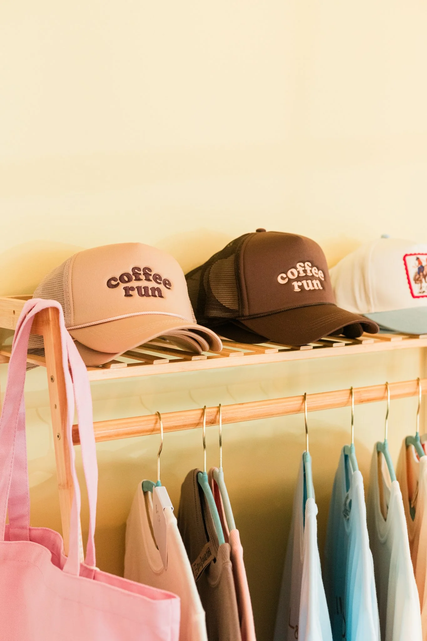 Three baseball caps on a shelf, two with the words 'coffee run' embroidered on the front, and a row of assorted clothing hanging below.