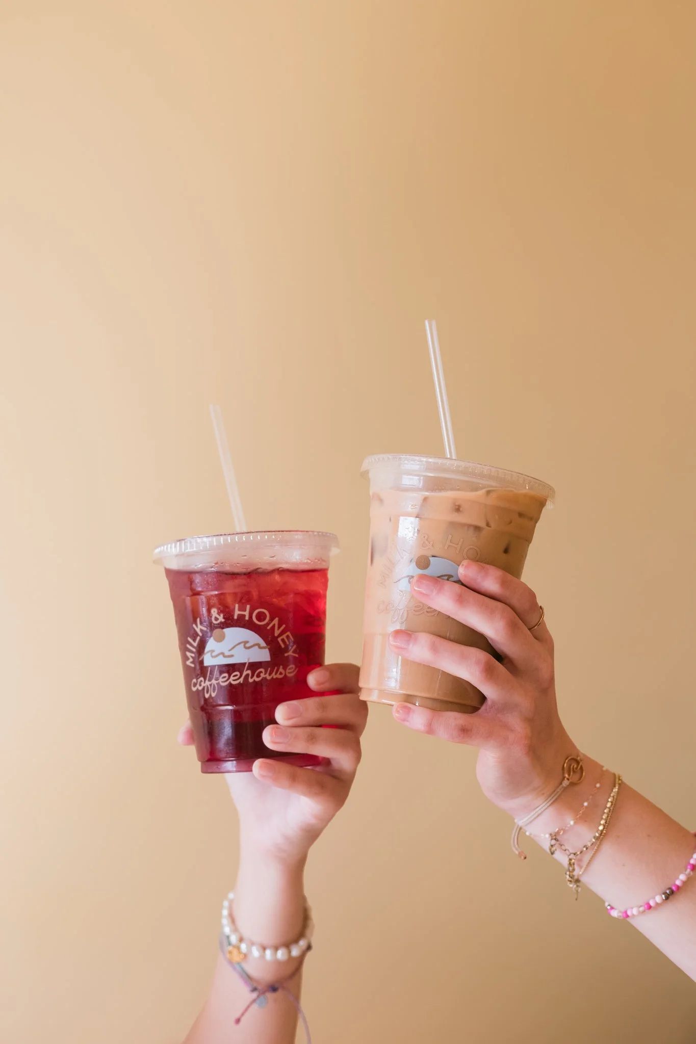 Two hands holding iced drinks in clear cups with straws, one with red liquid and the other with coffee-colored liquid, in front of a beige background.