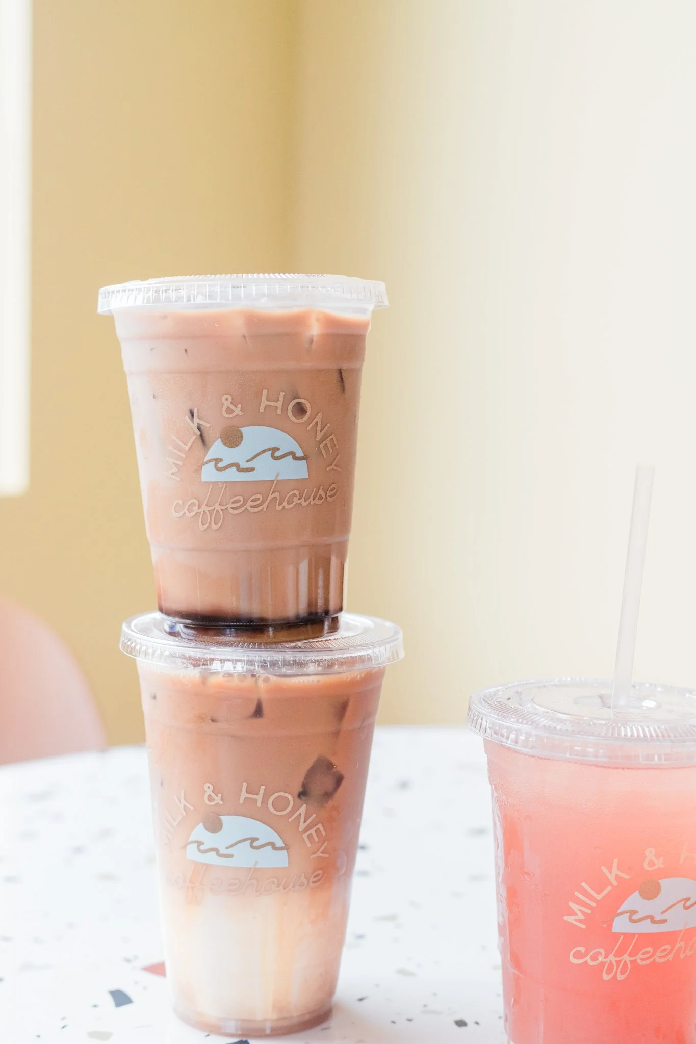 Two stacked plastic cups containing pink beverages with black tapioca pearls, and one additional pink beverage with a straw, all on a white speckled table.