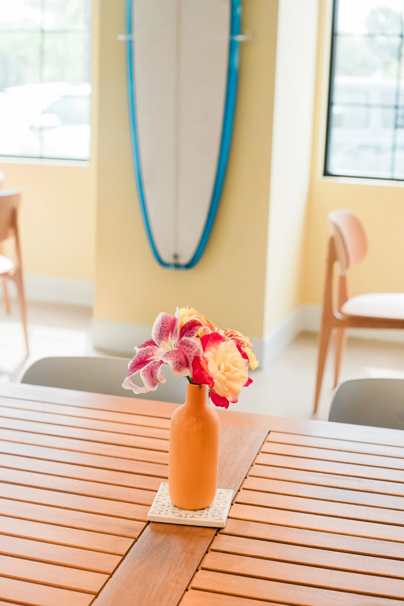 A small orange vase with pink and cream flowers on a wooden table in a bright room with yellow walls and large windows.