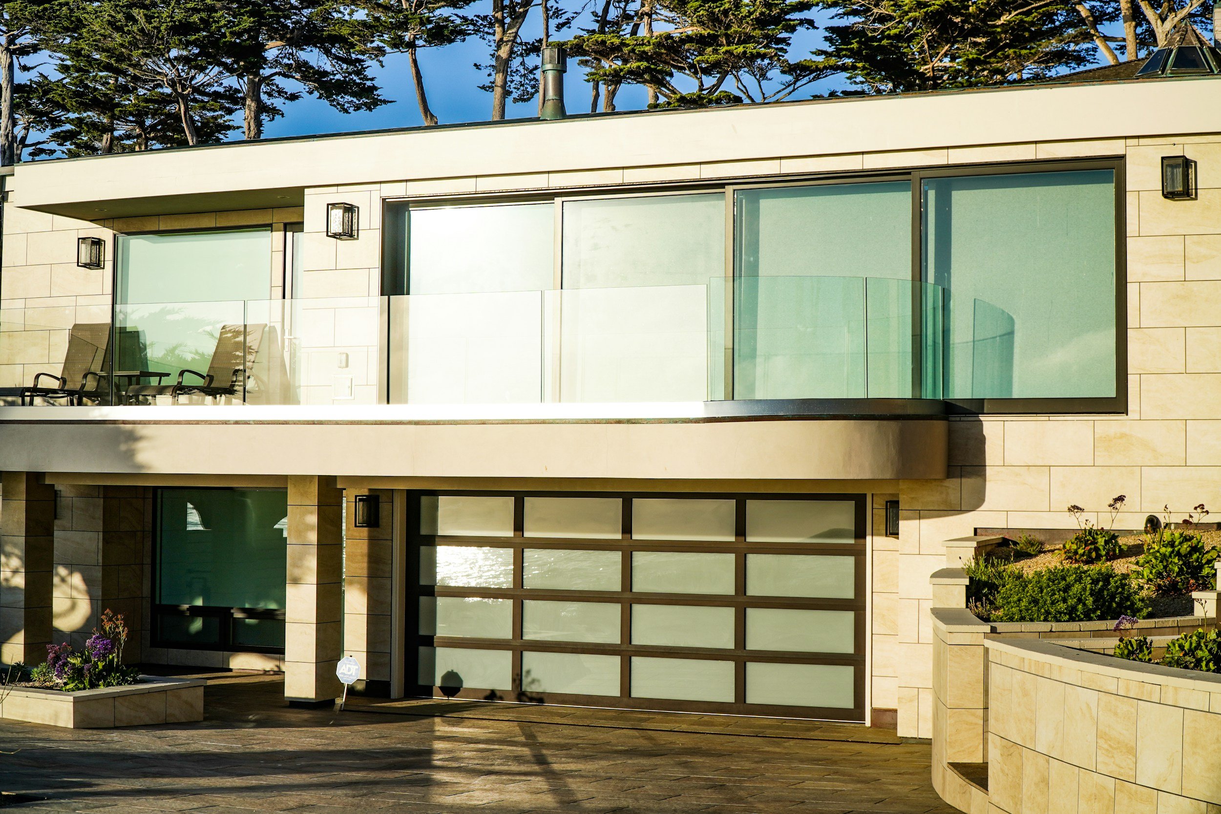 Modern two-story house with beige stone exterior, large glass windows, a glass balcony on the second floor, and potted plants, with trees in the background.