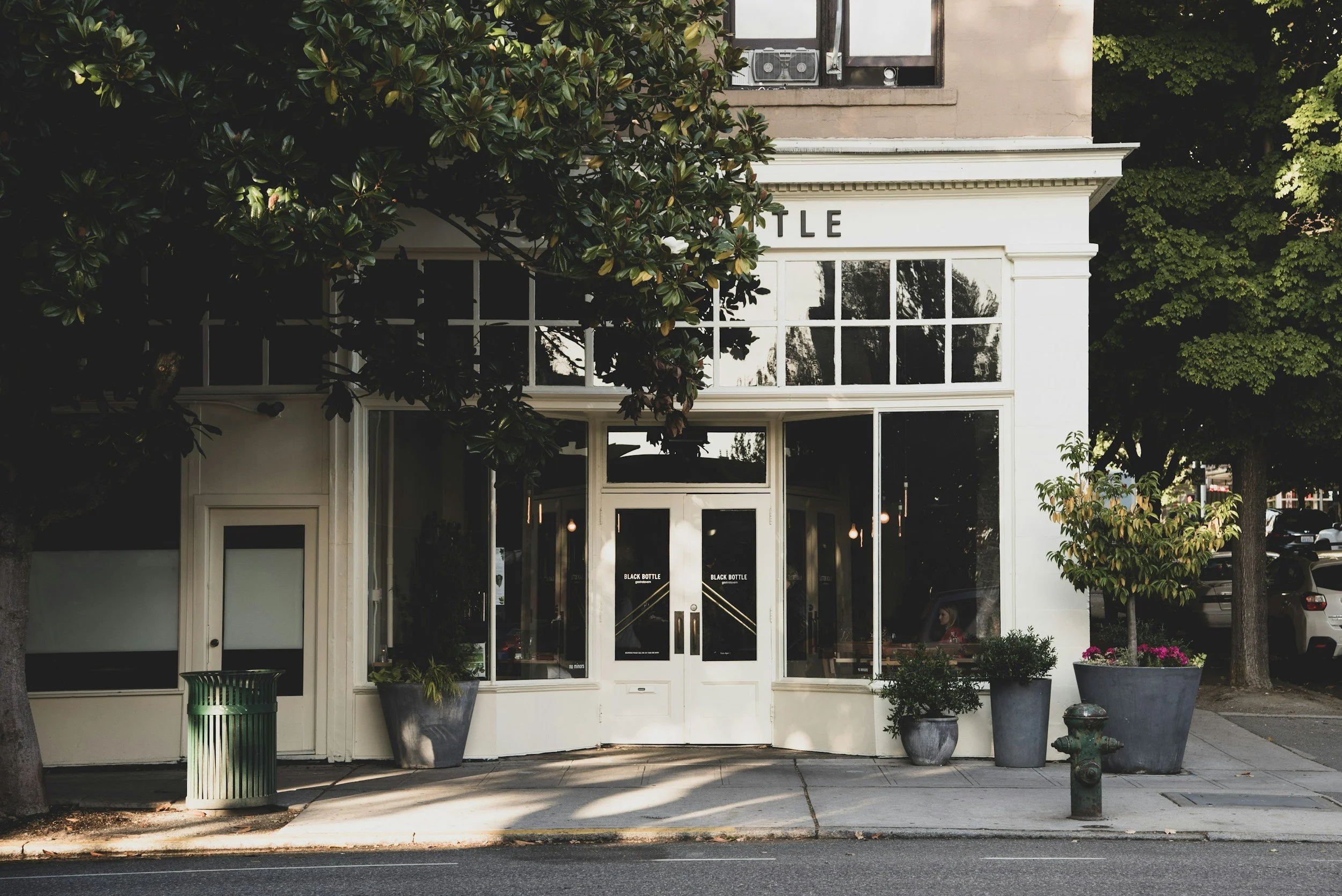 A storefront with a large glass window and door, displaying the words 'Black Bottle' on the door. The building is flanked by potted plants and is partially shaded by trees. The sidewalk in front is visible, with a green trash can on the left and a fire hydrant on the right.