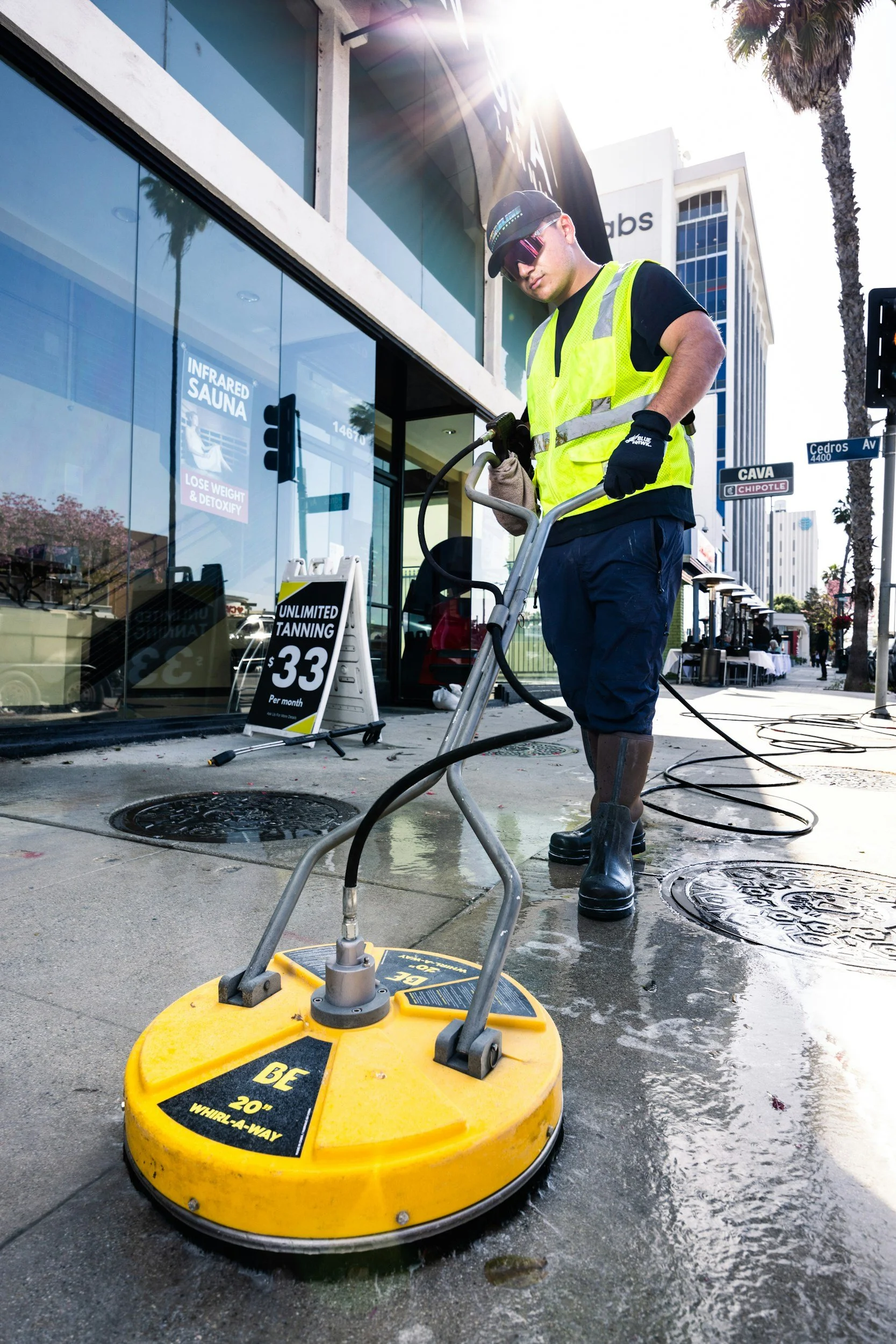 A man wearing a yellow safety vest, black gloves, and boots, using a yellow power washing machine on a sidewalk in an urban area with tall buildings and palm trees, during daytime.