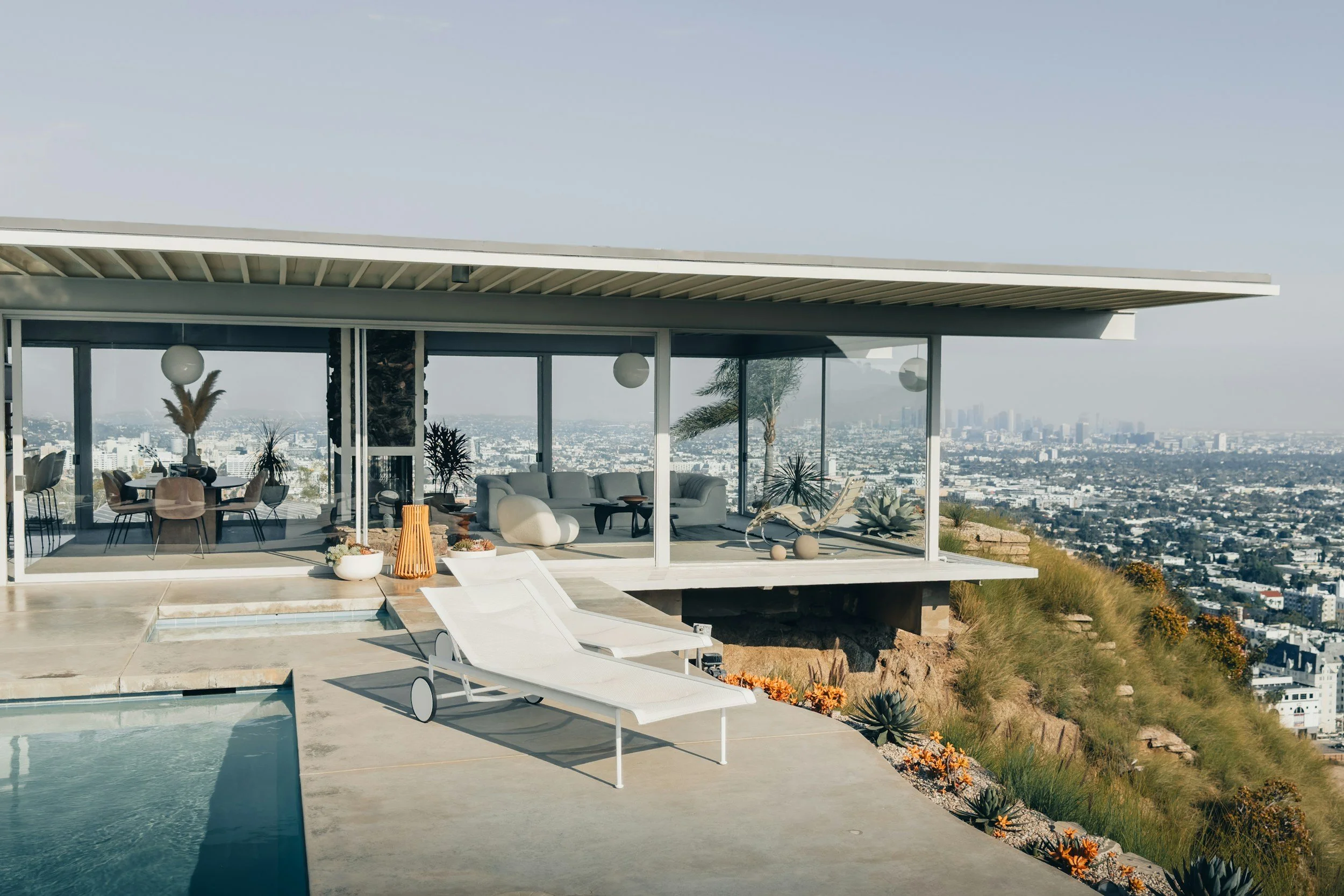 Modern house with large glass windows, outdoor pool, two white lounge chairs, and city view with buildings in the distance.