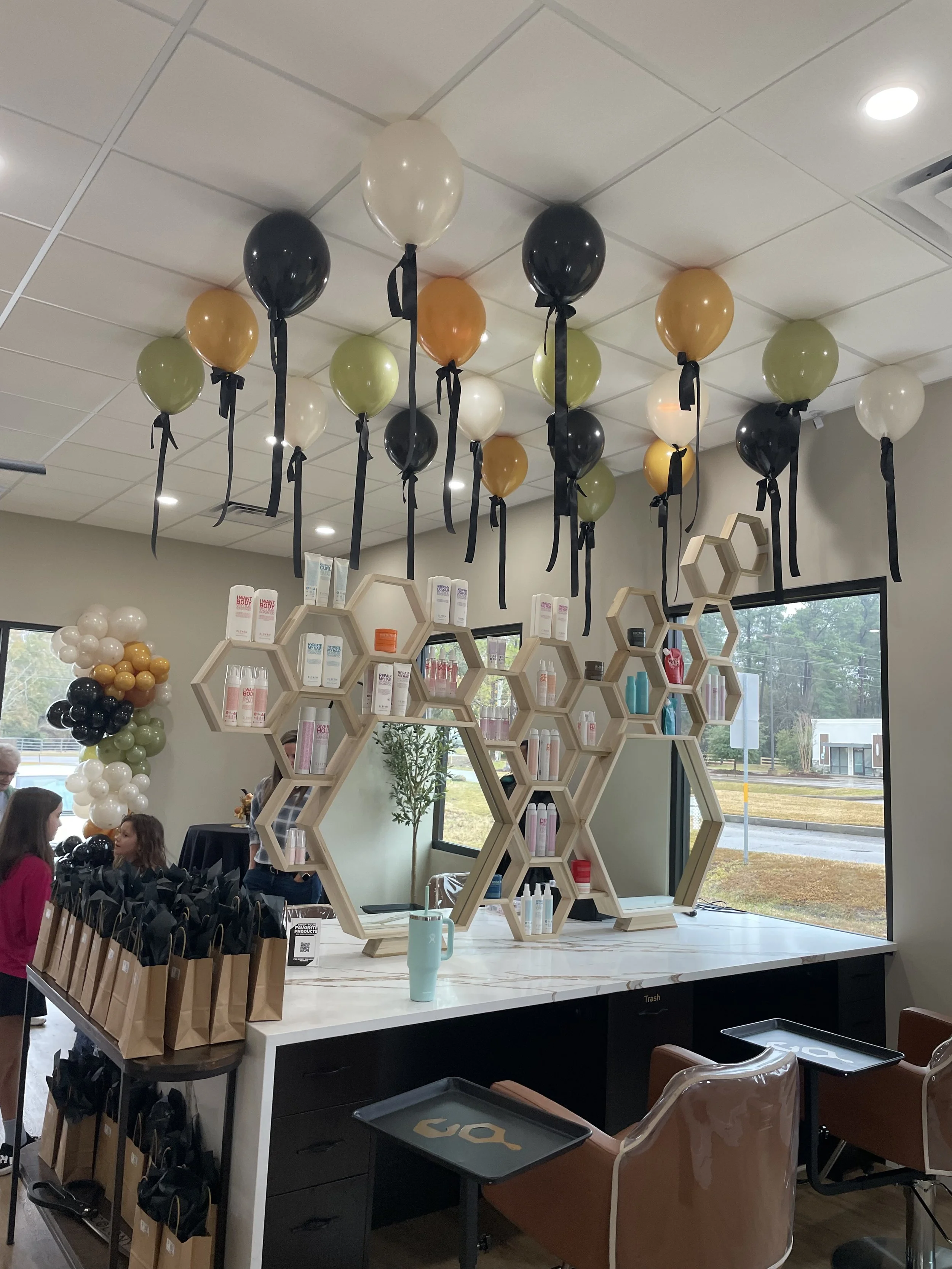 Decorated room with black, white, gold, and green balloons hanging from the ceiling, a marble countertop with paper bag gift bags, honeycomb shelves with products, and a large window showing an outside street view.