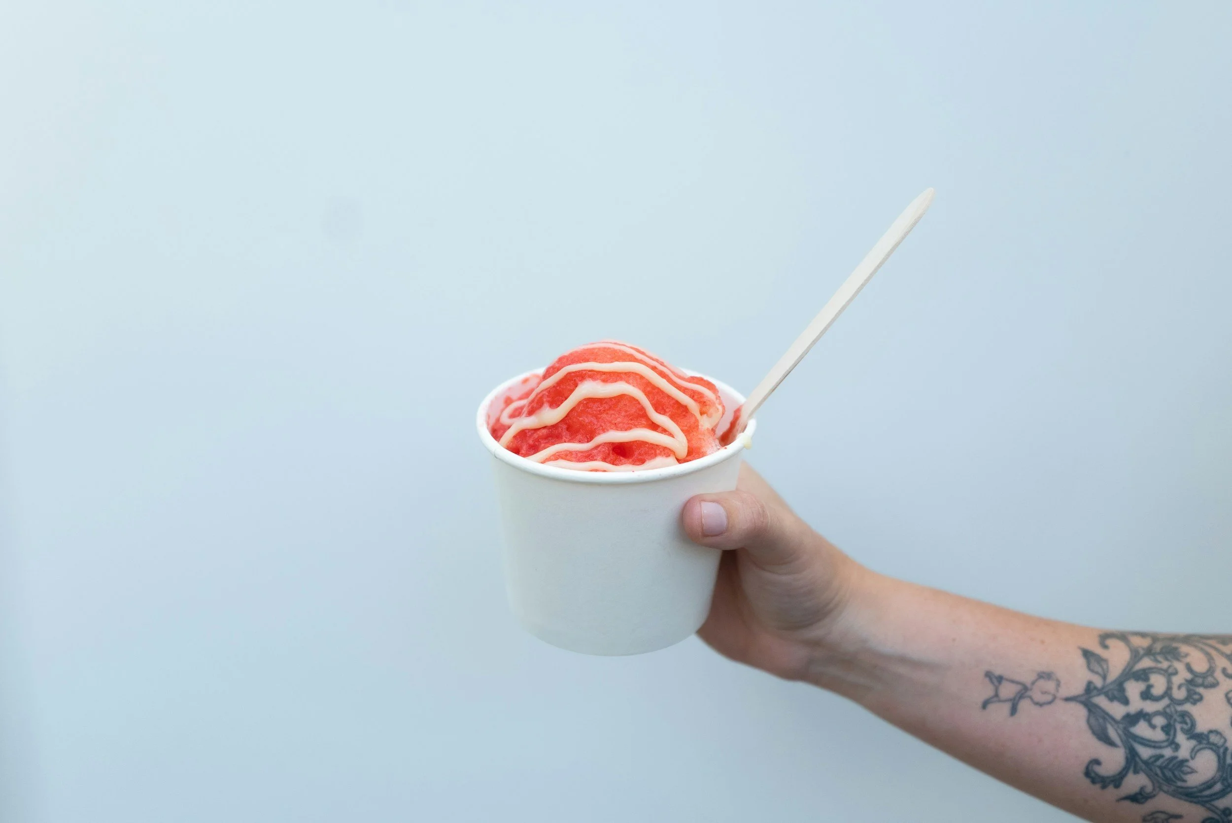 A person's hand with a tattoo holding a white paper cup filled with pink shaved ice drizzled with white syrup, and a white spoon protruding from the cup, against a plain light background.