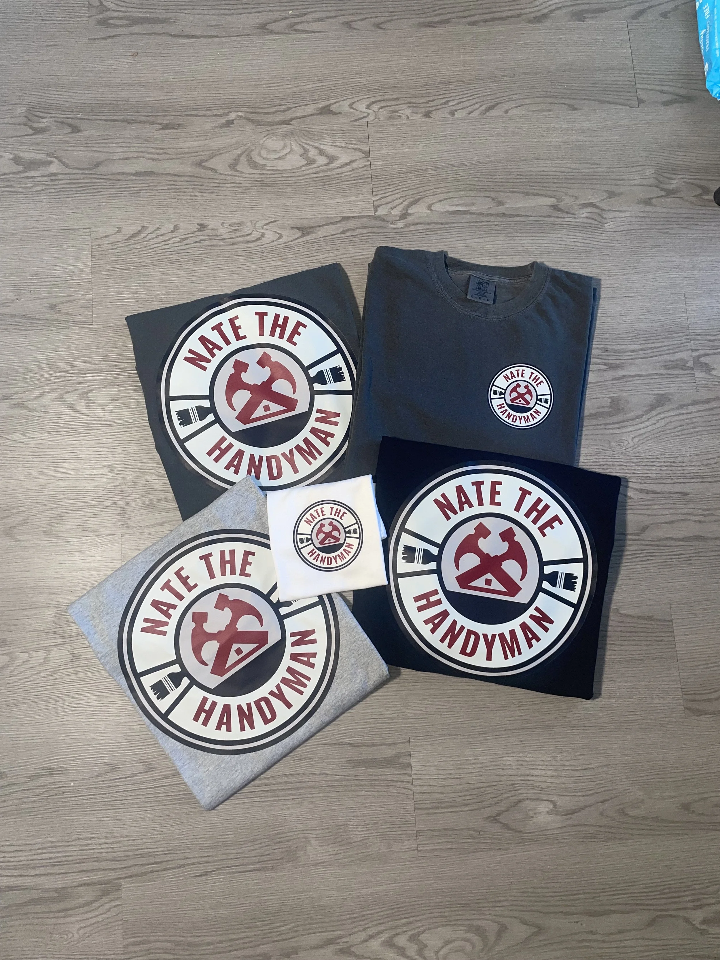 A collection of gray, black, and white t-shirts and a small sticker on a wooden floor, all featuring a circular logo with a red hammer and wrench crossed over each other, and the text 'Nate the Handyman' around the circle.