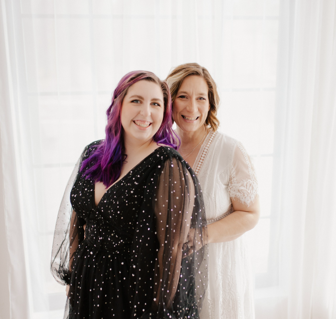 Two women smiling, standing in front of a window with white curtains. One has purple hair and is wearing a black dress with white sparkles. The other has brown hair and is wearing a white lace dress.