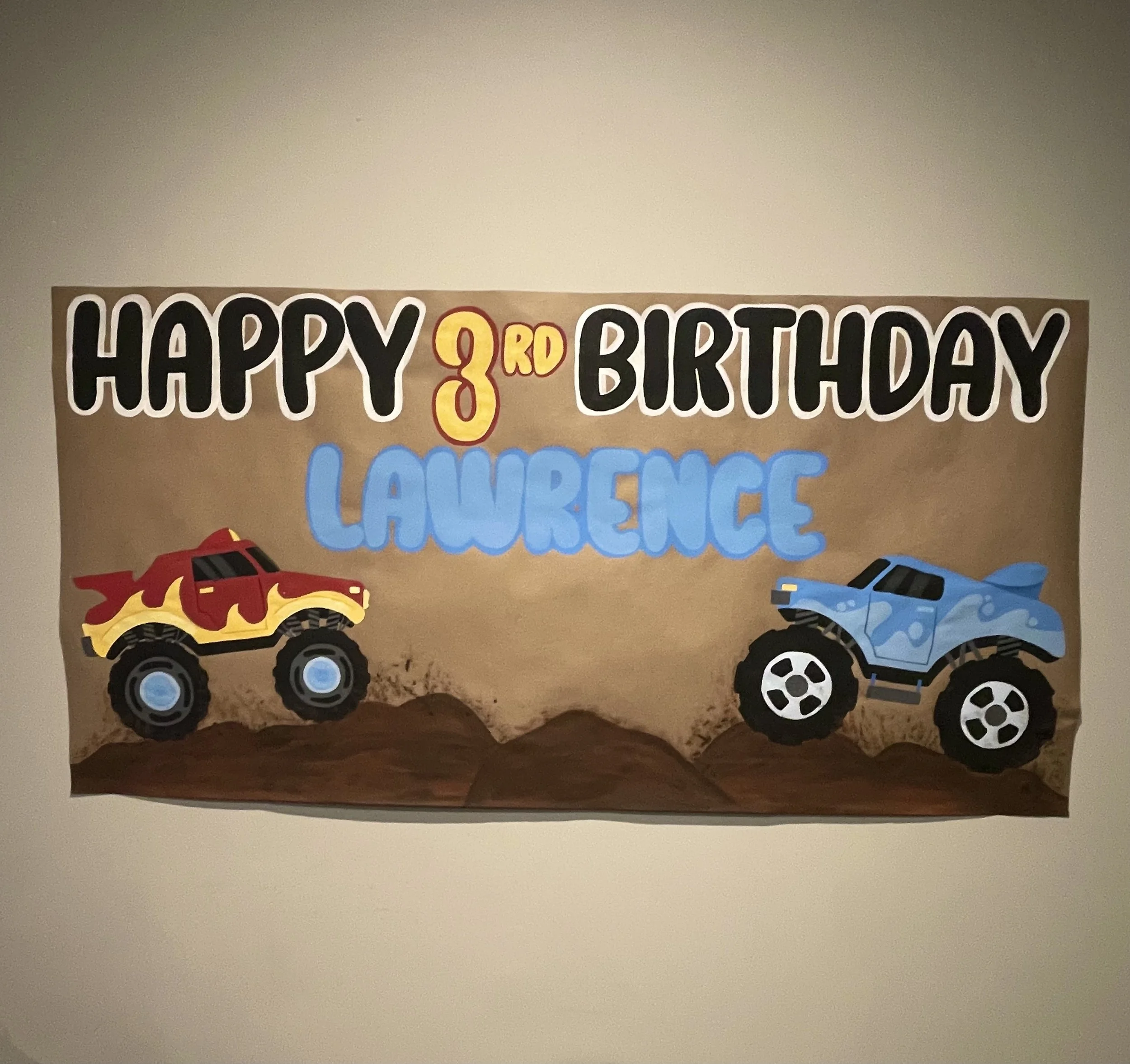 Decorative birthday poster with toy monster trucks, a red and yellow one on the left and a blue one on the right, celebrating Lawrence's third birthday.