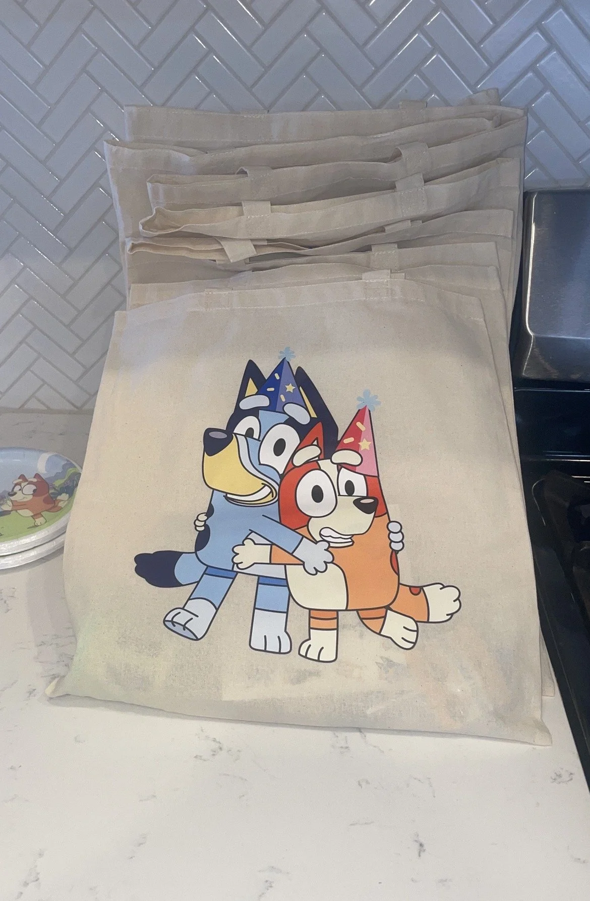 A beige tote bag featuring a graphic of two cartoon dogs wearing colorful birthday hats, hugging each other, on a white kitchen countertop.