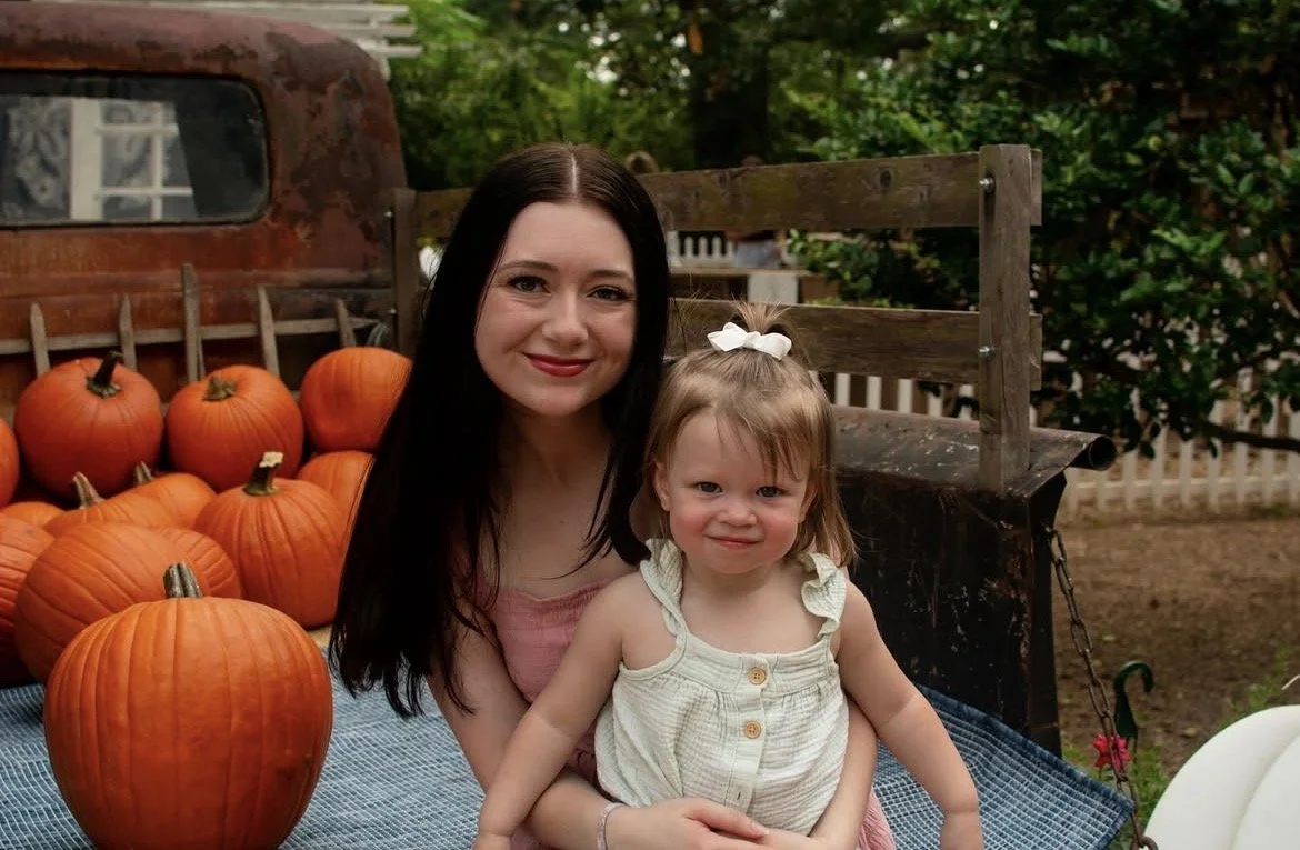 A woman with long dark hair and a young girl with light hair and a white bow, smiling for the camera, outdoors near pumpkins on a truck bed.