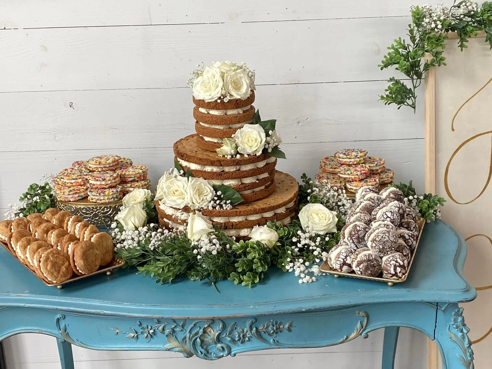 A dessert table with a tiered cake made of cookies and cream, decorated with white roses and greenery, surrounded by cookies and other treats on a blue vintage table against a white wooden background.